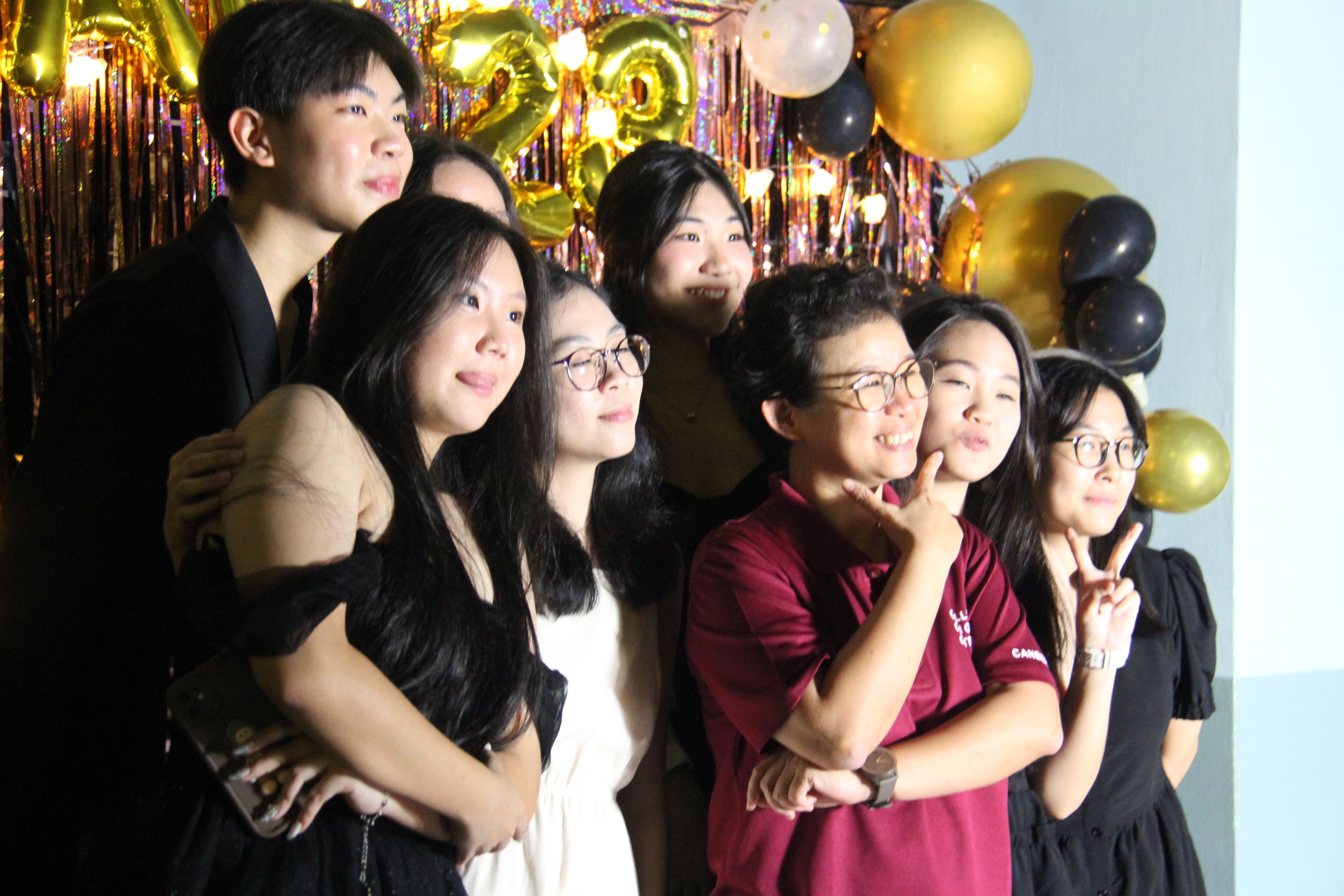 Group of eight young people posing at a party with gold and black balloons and a shiny gold backdrop.