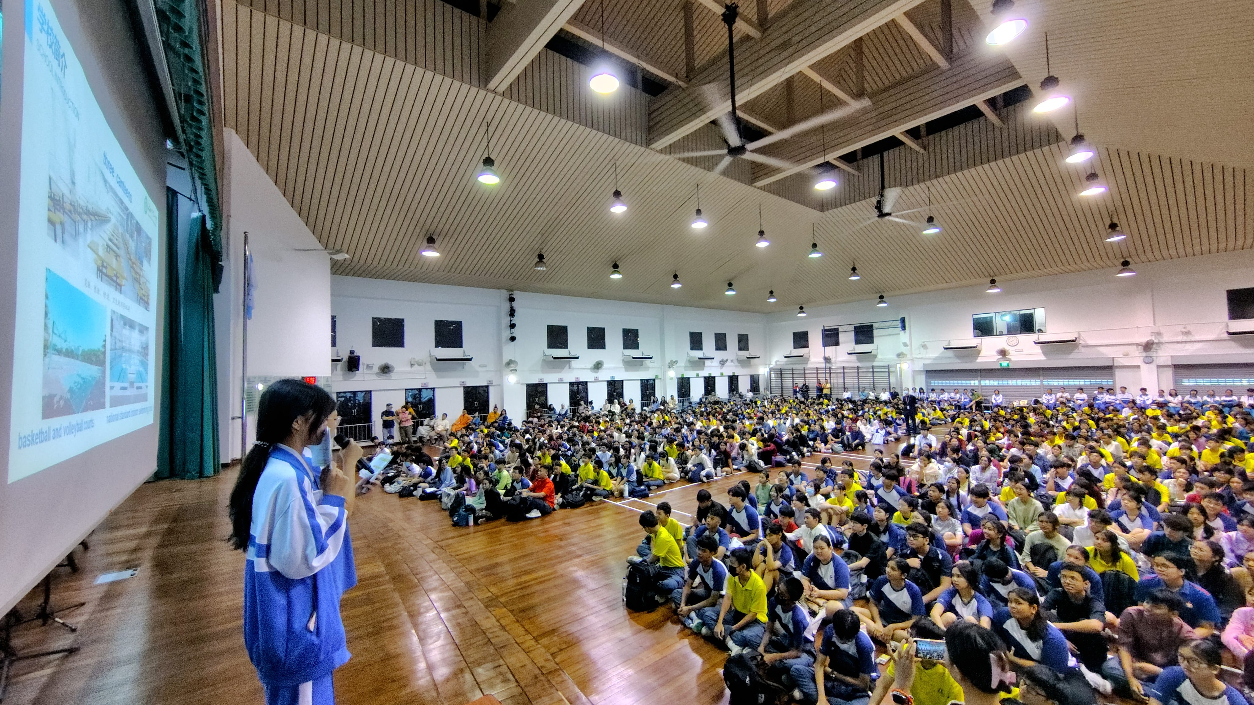 A student speaks into a microphone before hundreds of students seated on a gym floor, with a presentation projected on screen.