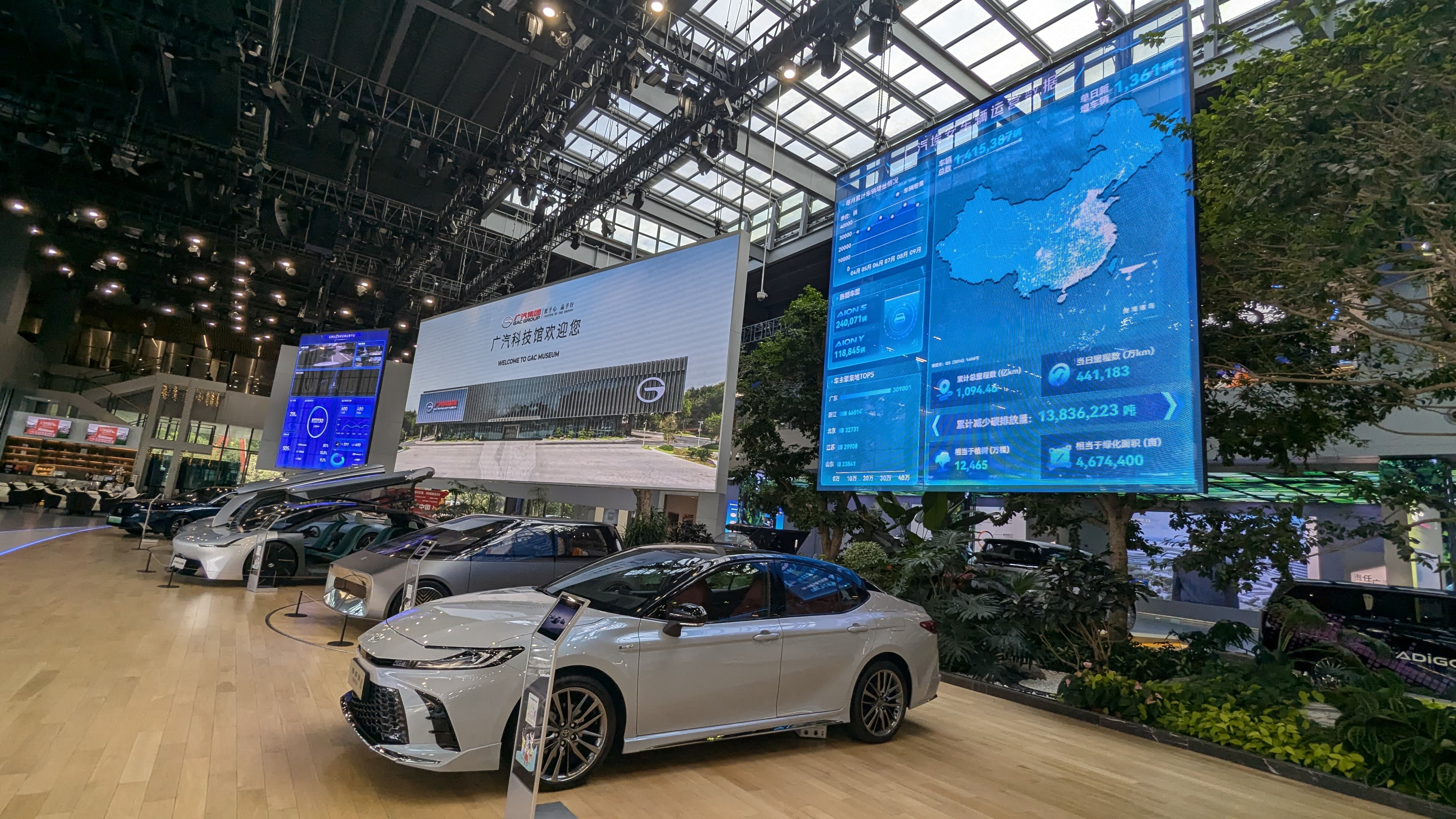 GAC Museum showroom with several cars on a wood floor, large LED screens displaying Chinese text and a map.