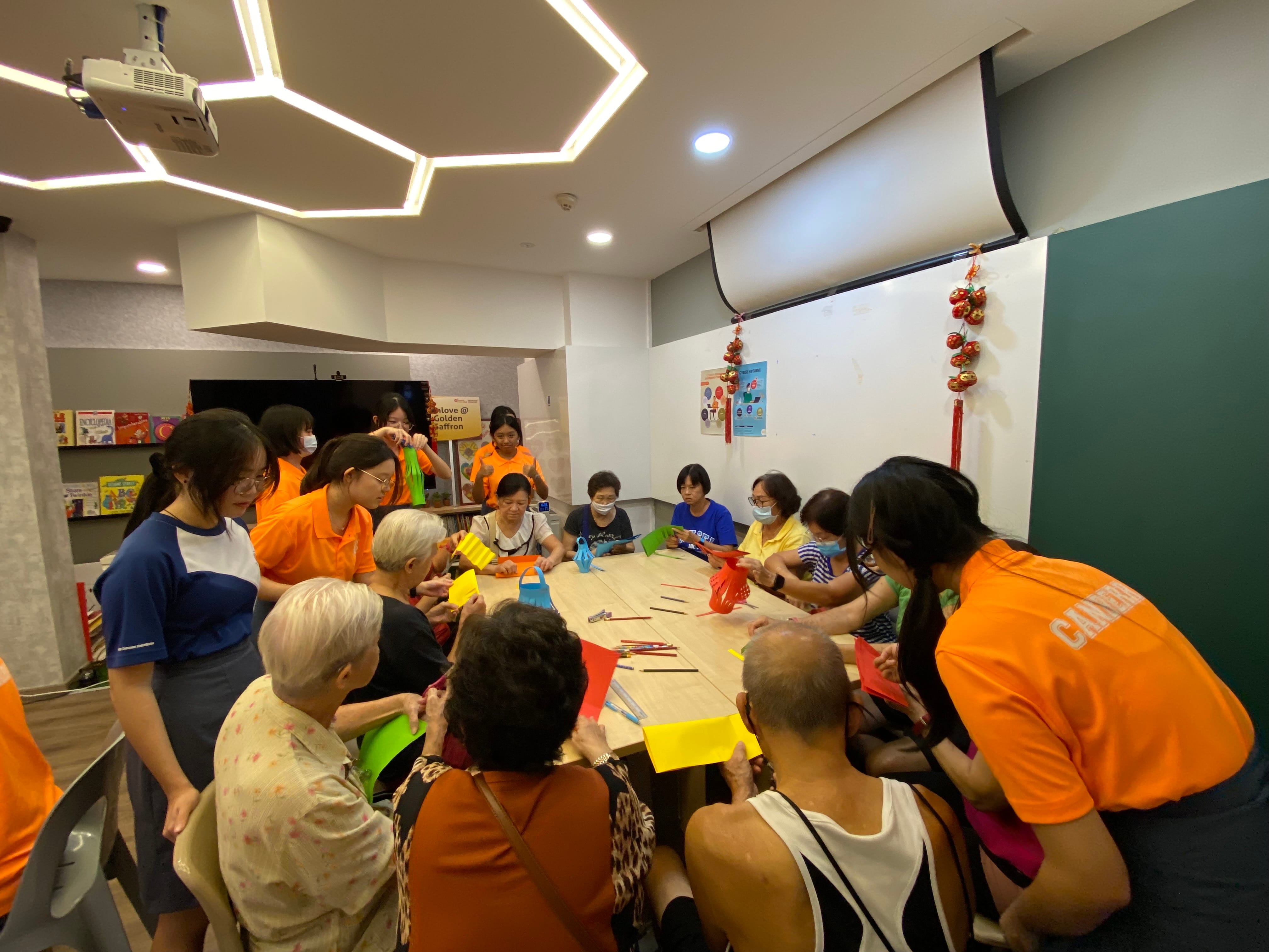 Students in orange tees gather around a table with senior citizens making paper lanterns