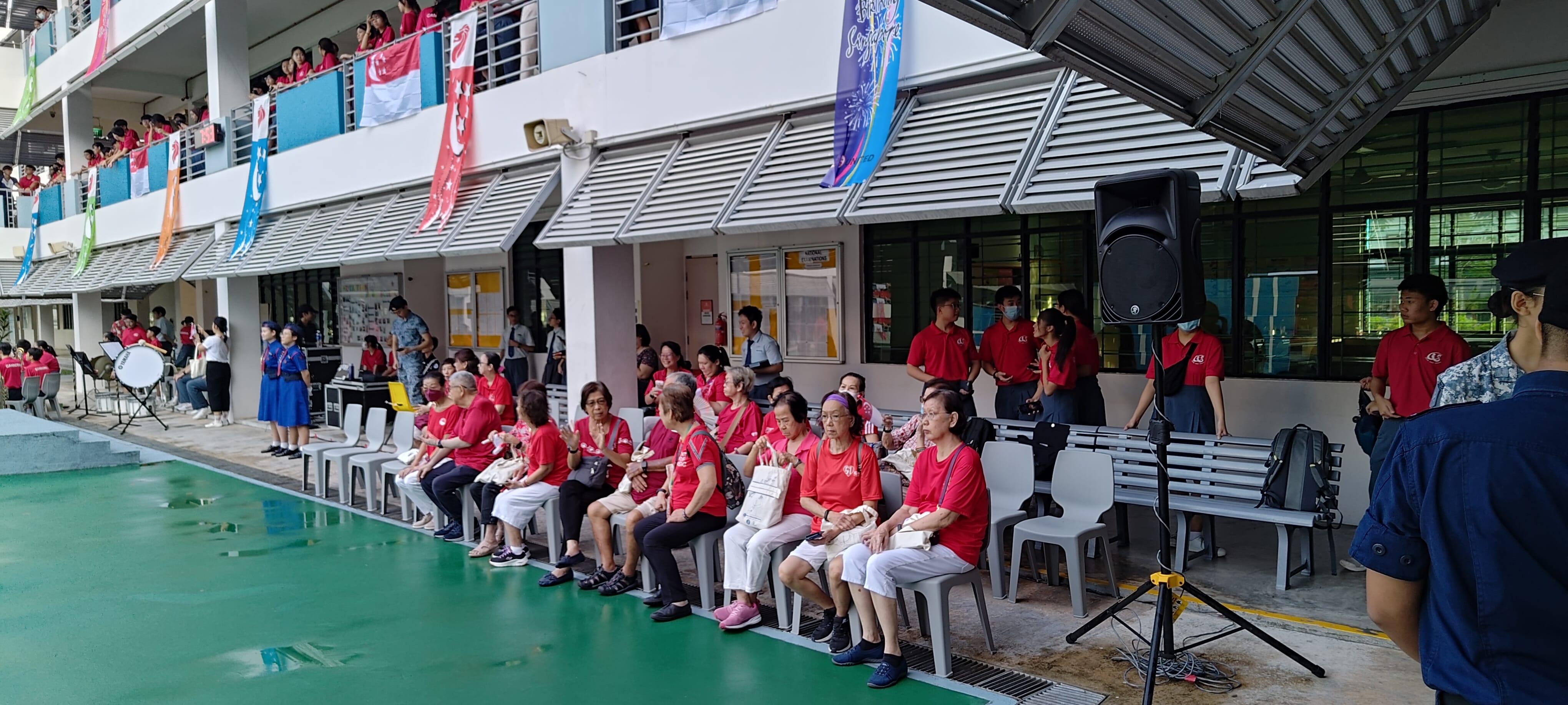 Elderly people in red shirts sit on chairs at an outdoor community event with colorful flags and a crowd nearby.