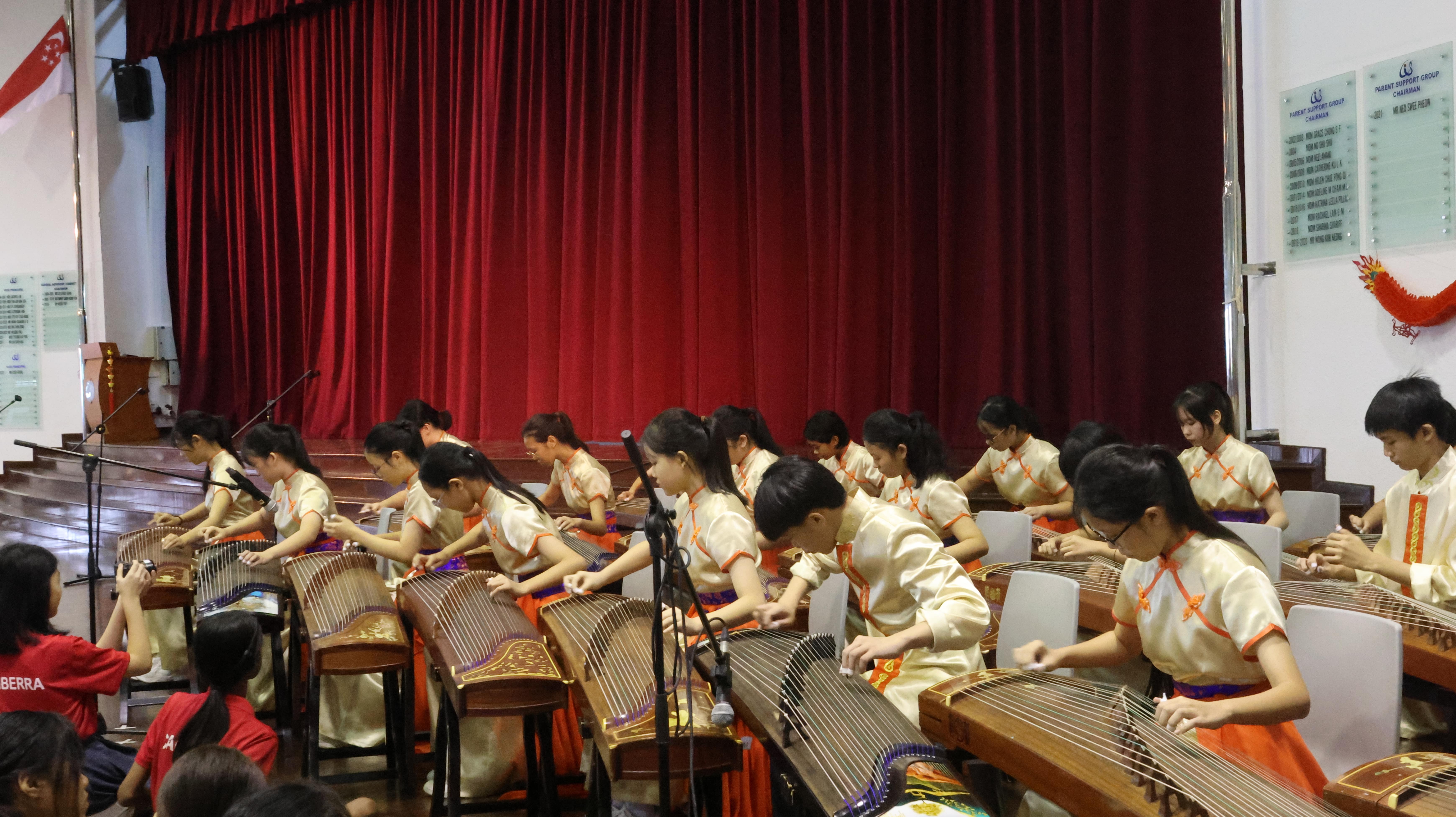 A group of young students in traditional Chinese attire playing guzheng instruments on a stage with red curtains.