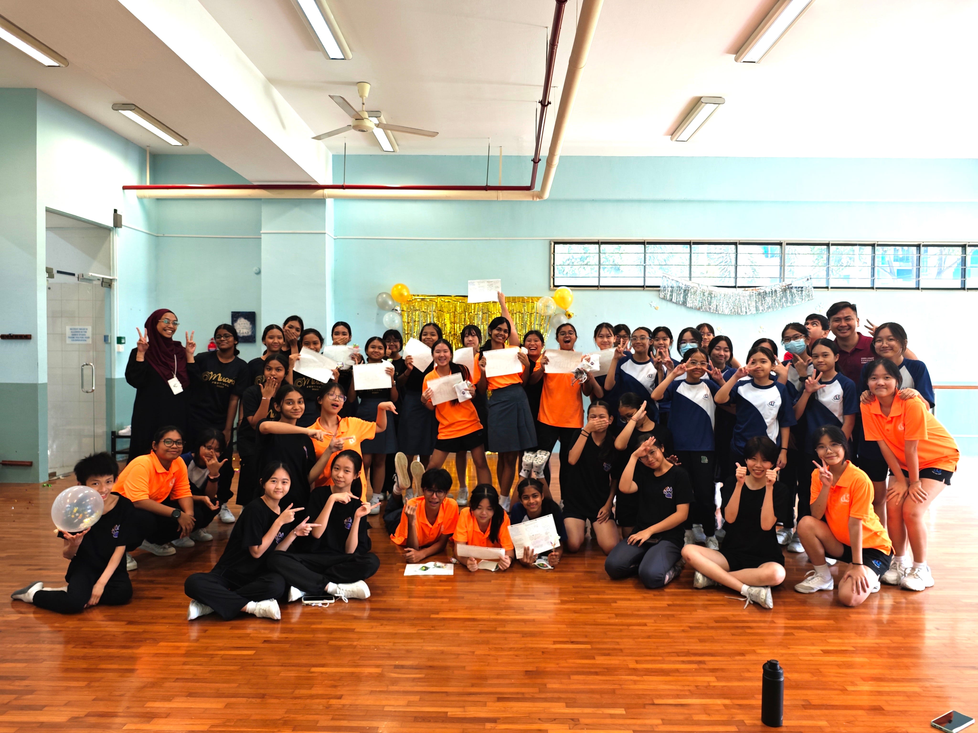 A large group of students in orange and dark uniforms pose together in a school hall with balloons and certificates.