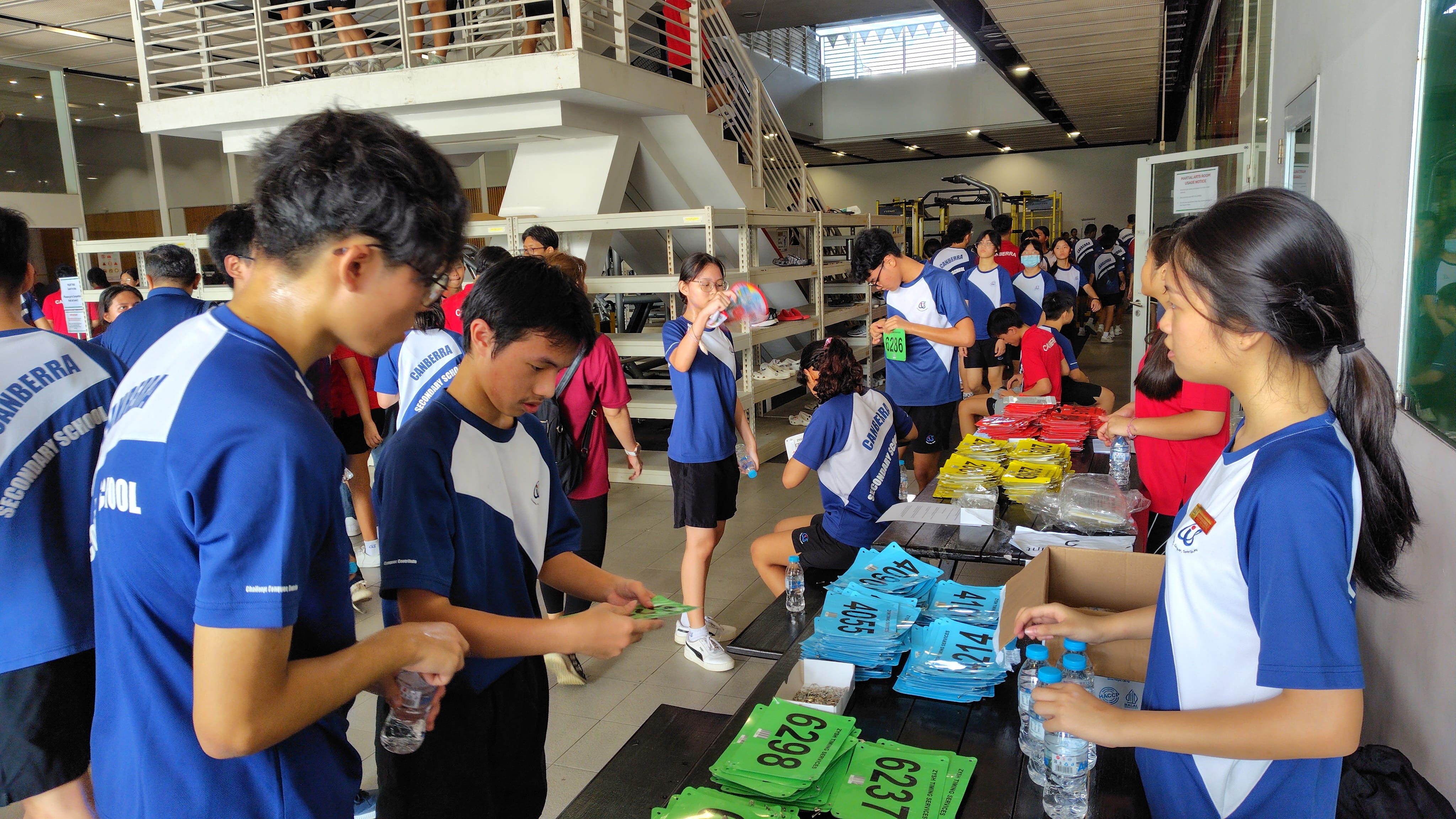 Students in Canberra Secondary School uniforms collect numbered race bibs in green, blue, and yellow at a registration table.
