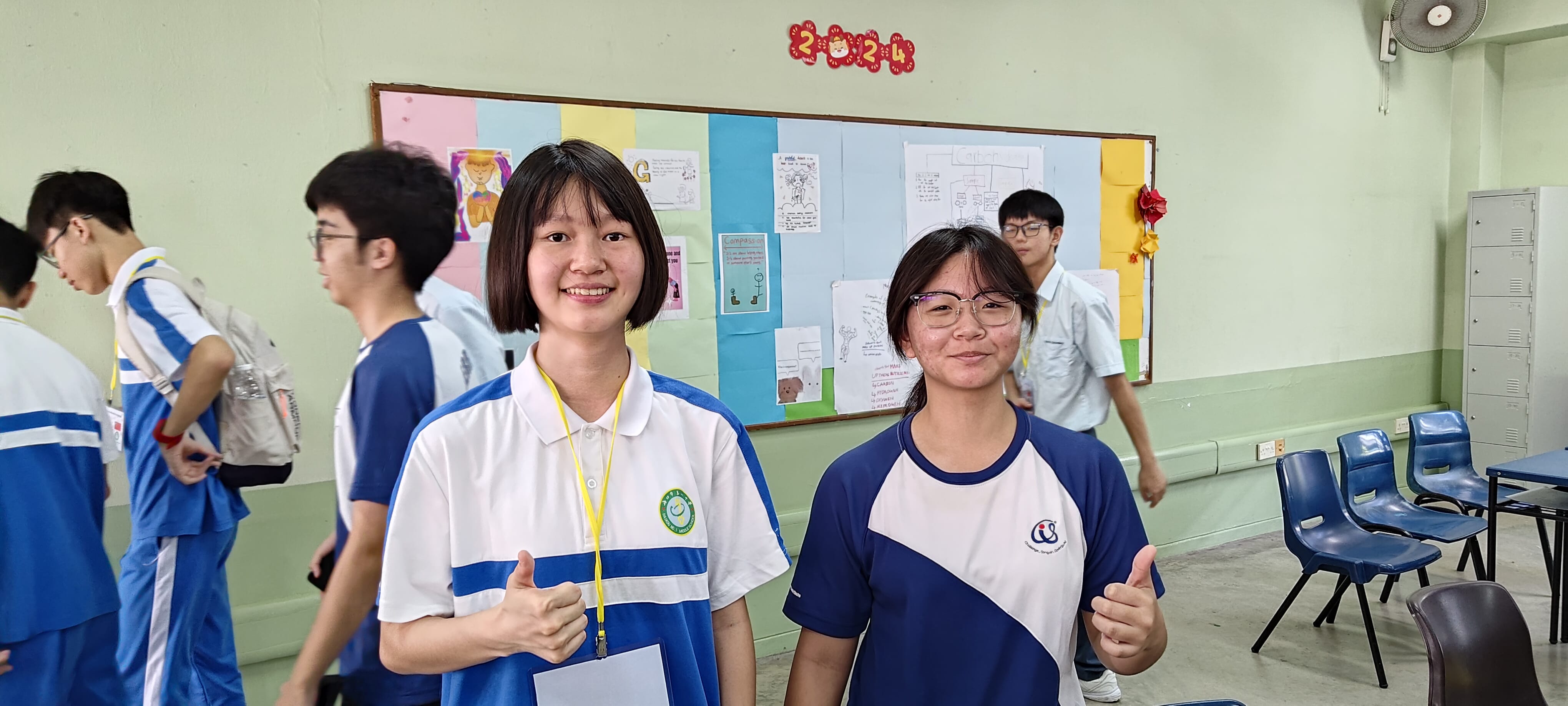 Two smiling female students in blue and white uniforms give thumbs up in a classroom with a colorful bulletin board.