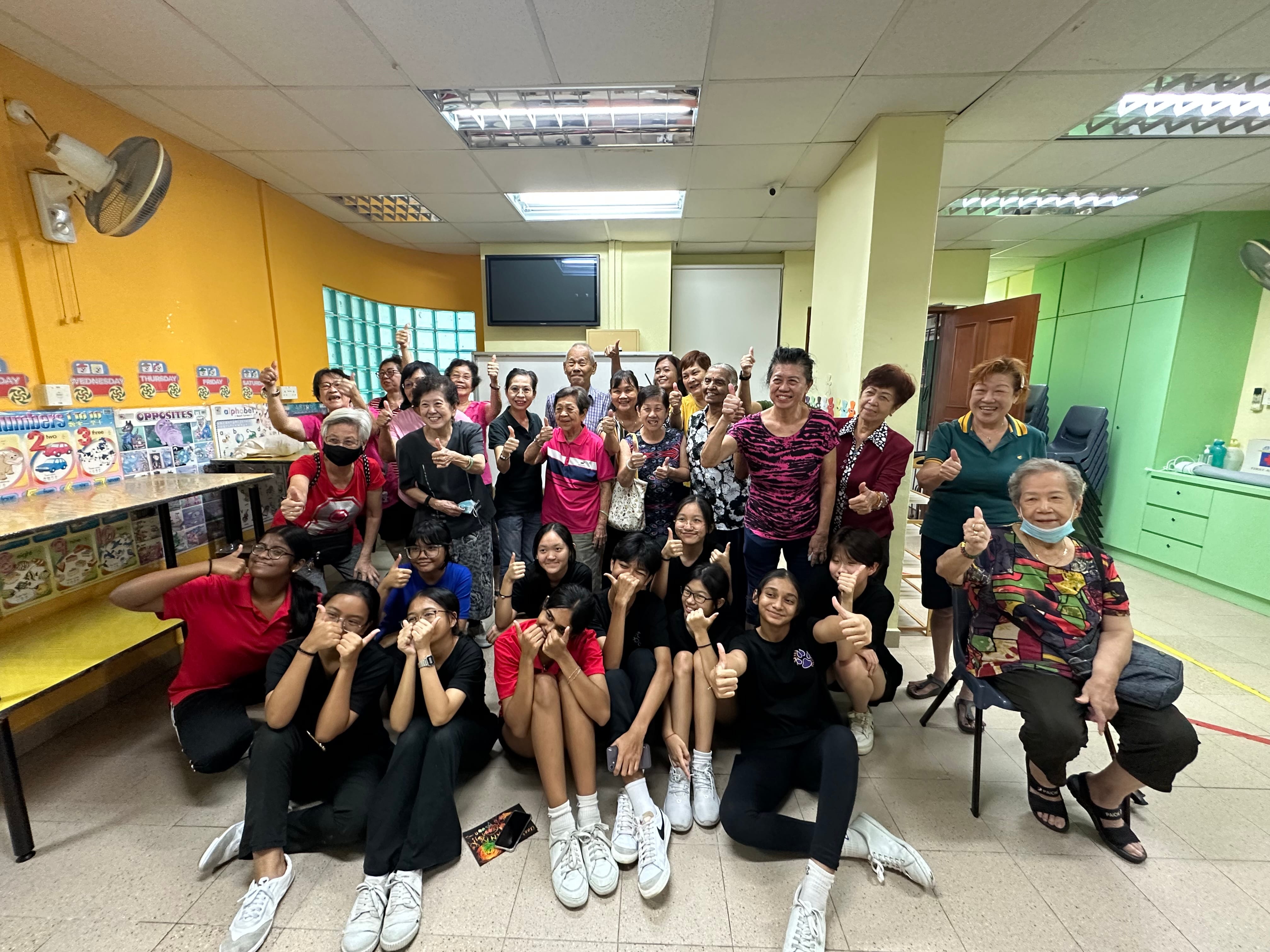 A large mixed-age group of people posing indoors with thumbs up, seated and standing in a colorful room.