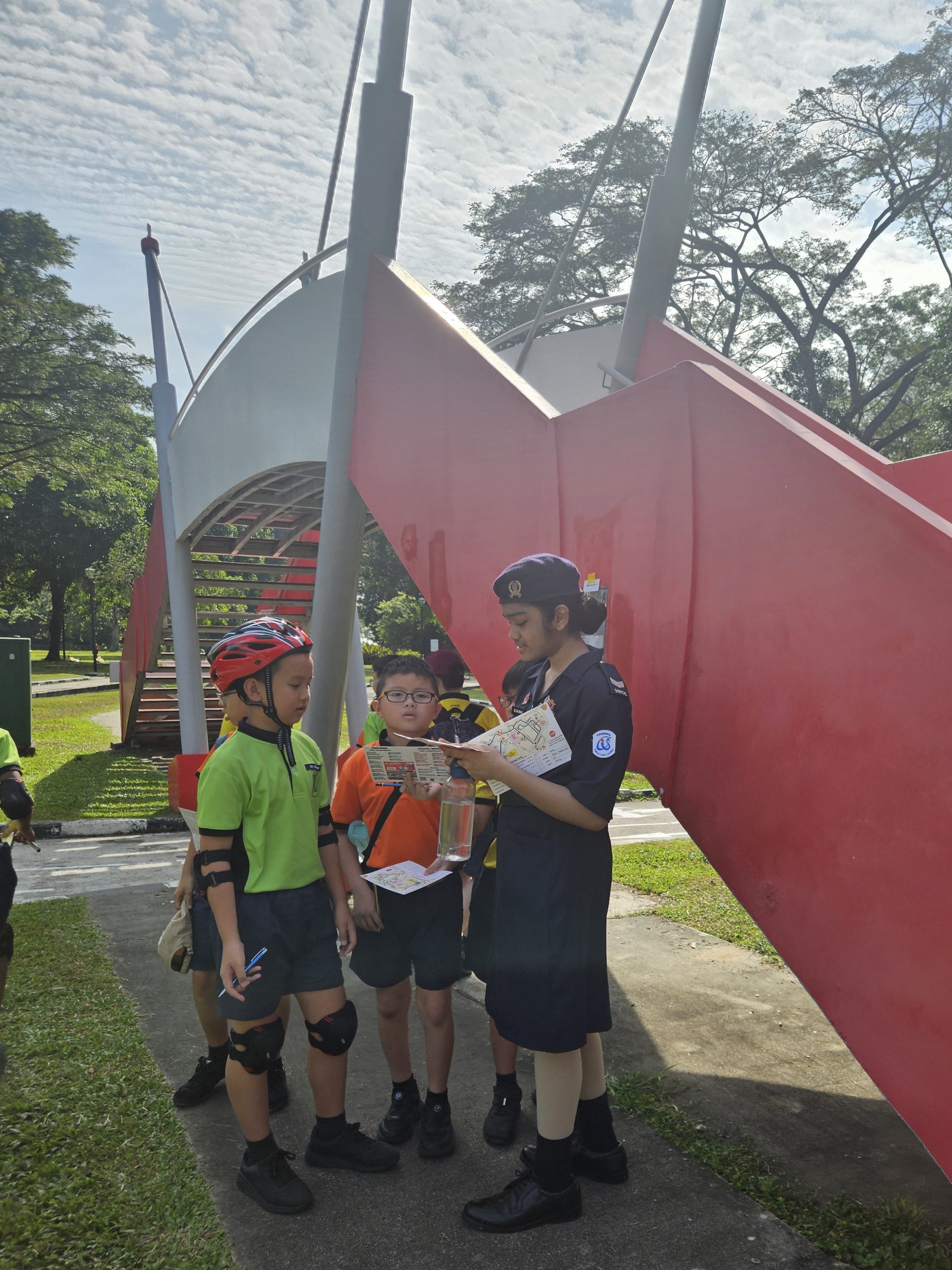 Three children in a park look at a map near a large red and white sculpture, one wearing a red helmet.