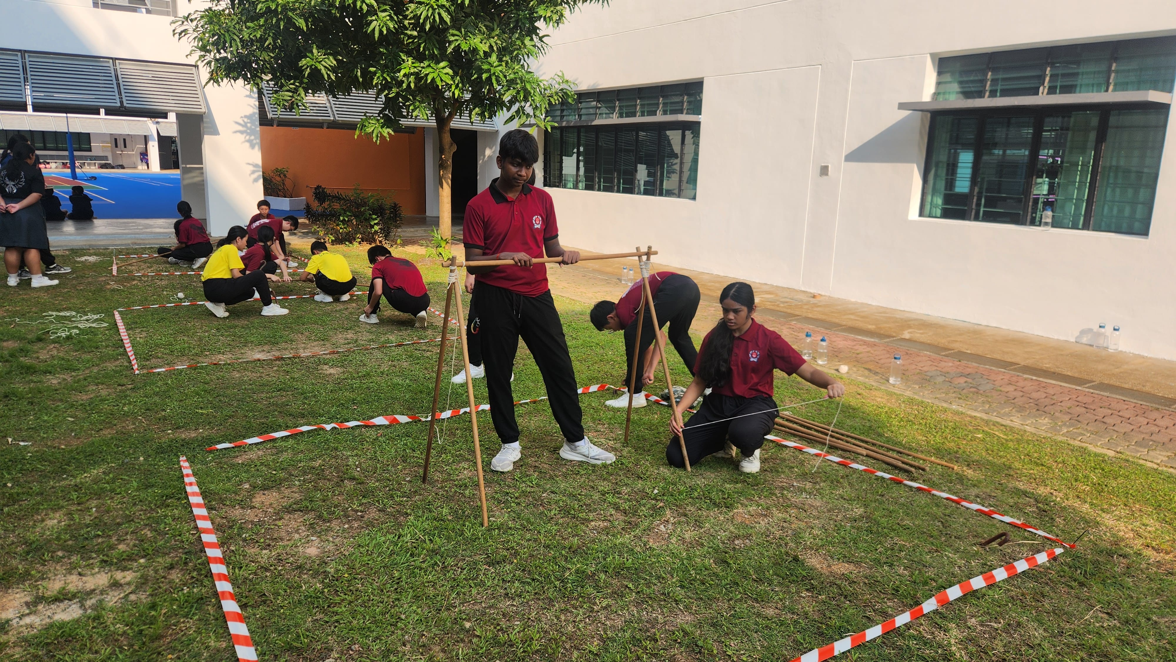 Students in red uniforms build wooden stick structures on grass within red-and-white tape boundaries outside a school.