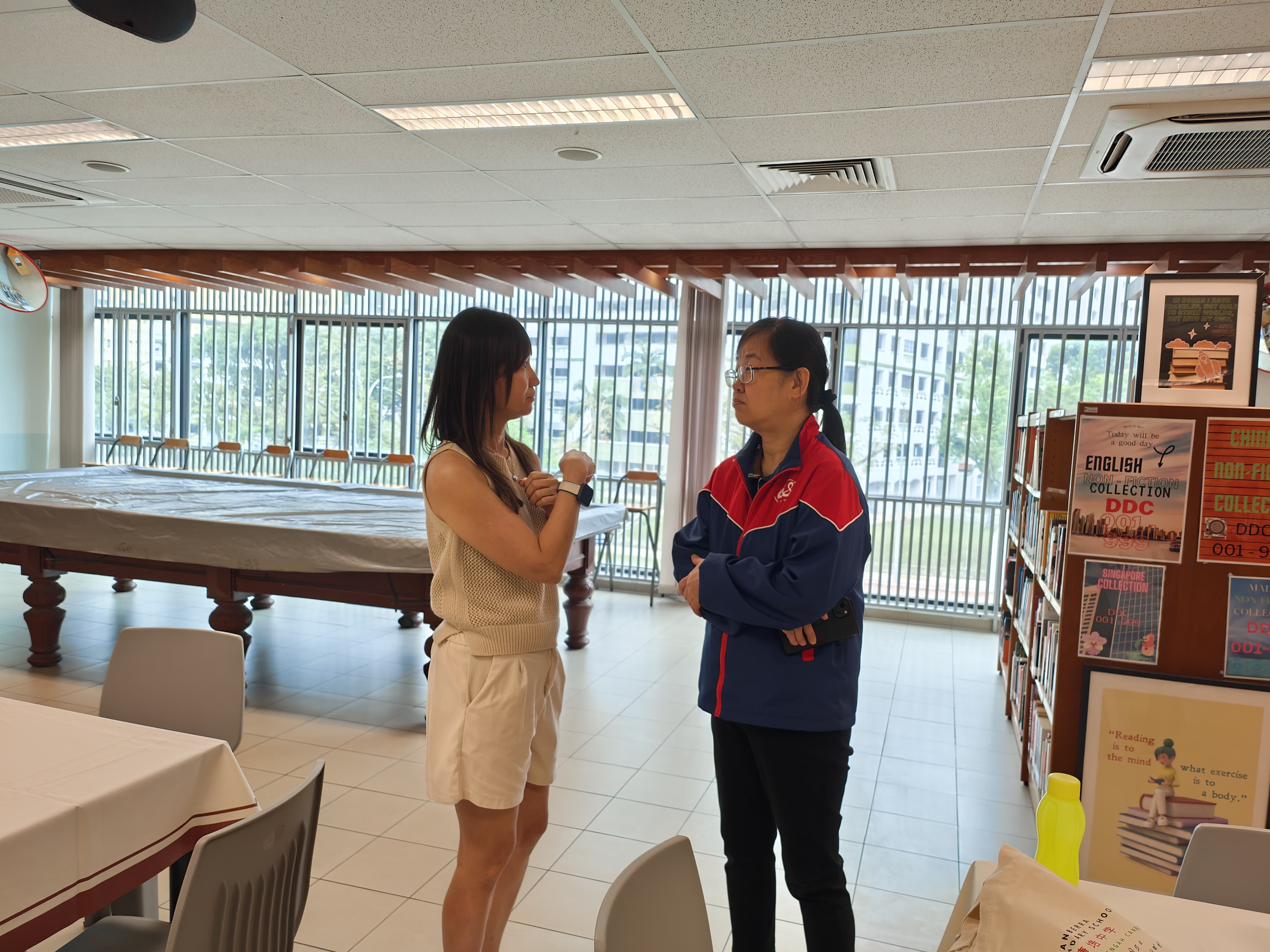 Two women conversing in a library with bookshelves, posters, and a covered billiard table in the background.