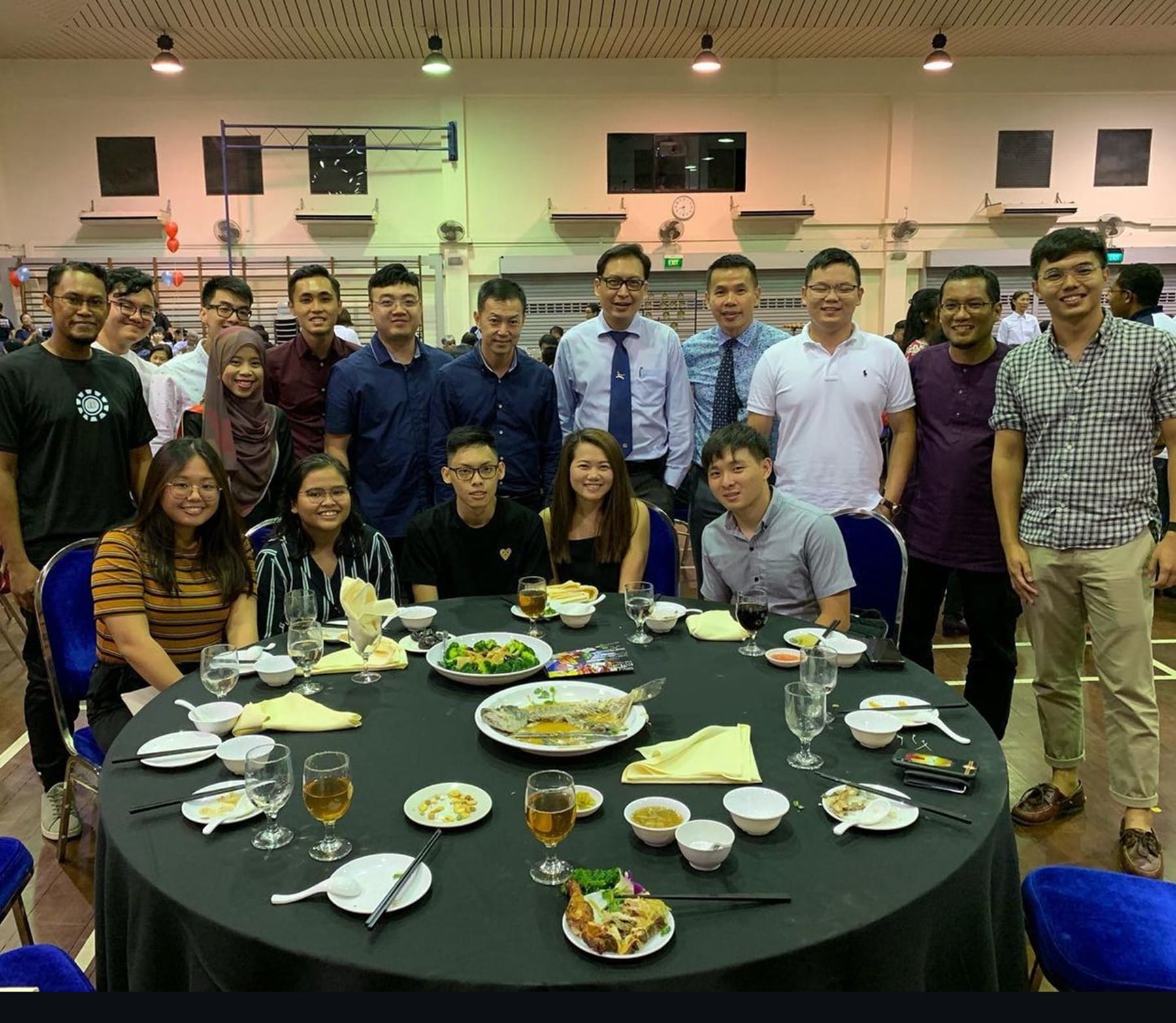 A large group of people pose around a round dining table with food and drinks at an indoor banquet event.