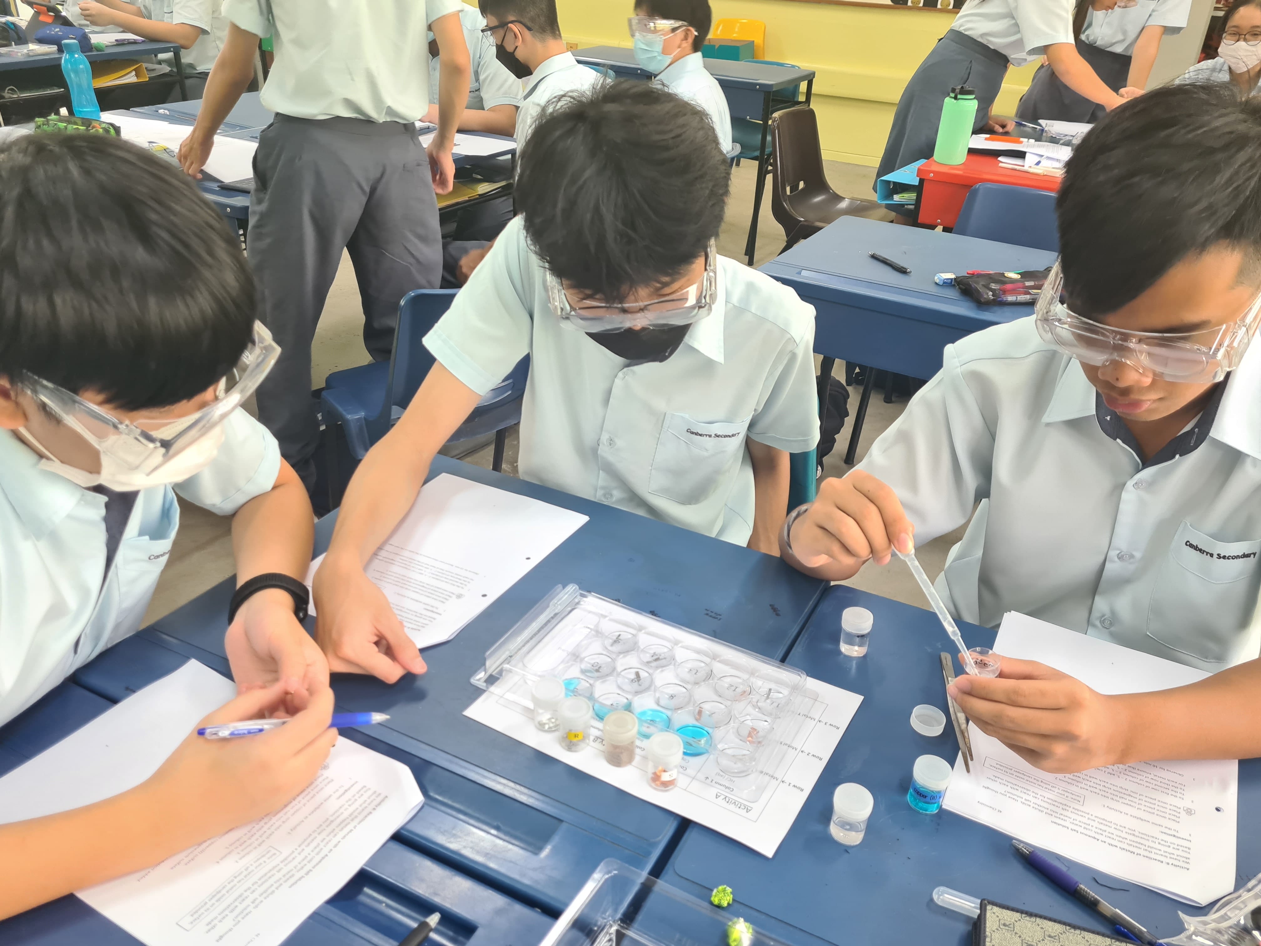 Three students wearing goggles and masks conduct a science experiment with small sample wells and a pipette at a blue desk.
