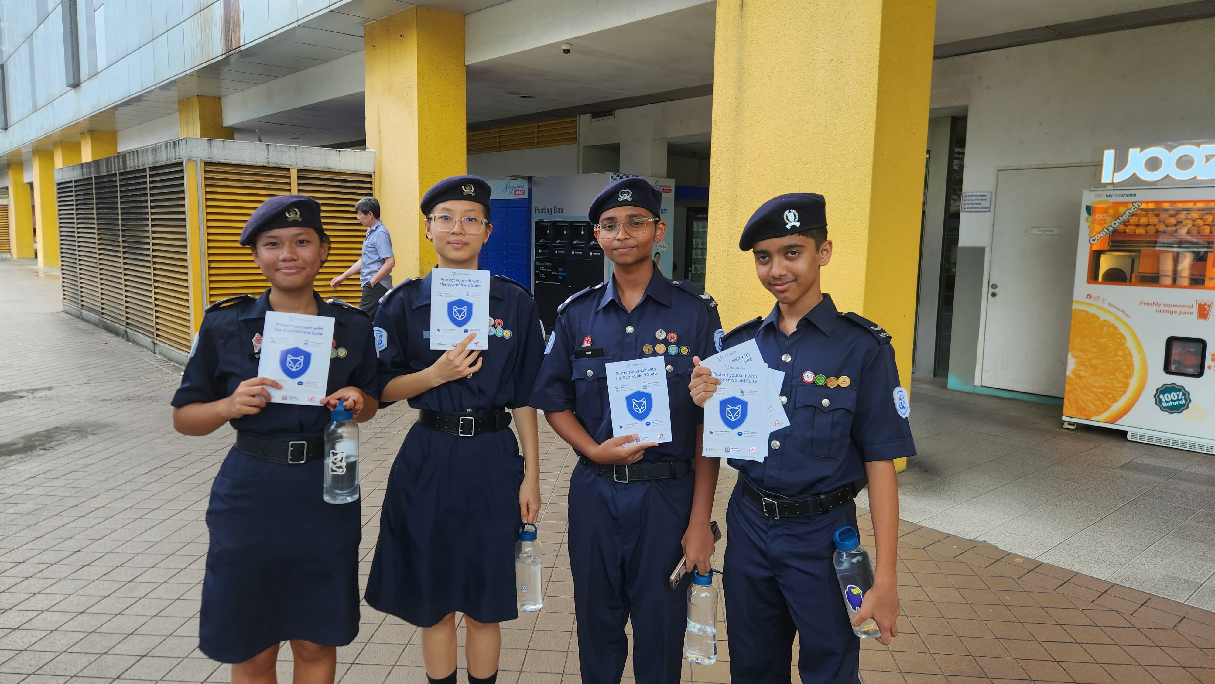 Four youth in navy blue uniforms and berets hold pamphlets outdoors near yellow pillars.