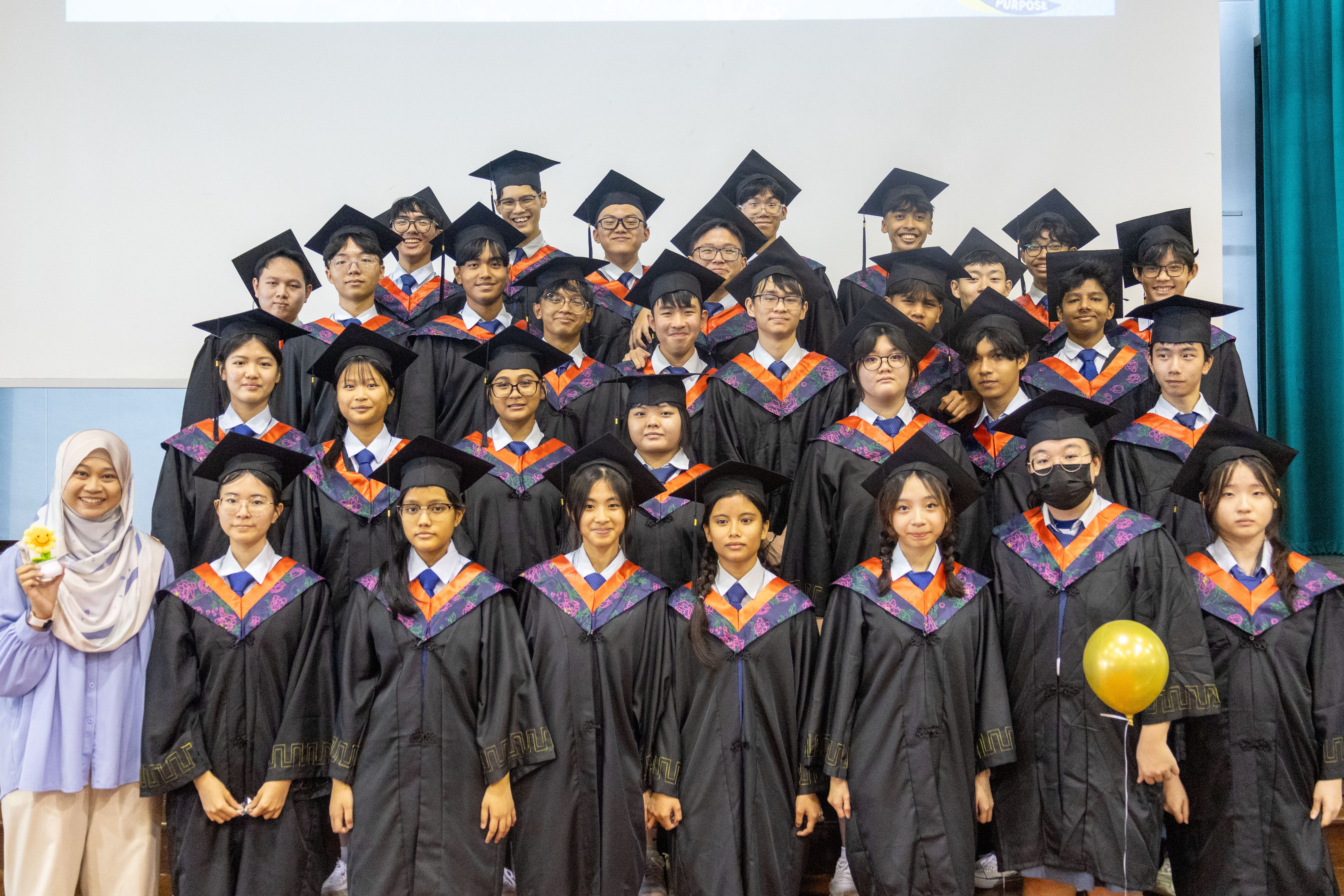 Group of graduates in black gowns and caps posing indoors; teacher at left holds a small trophy.
