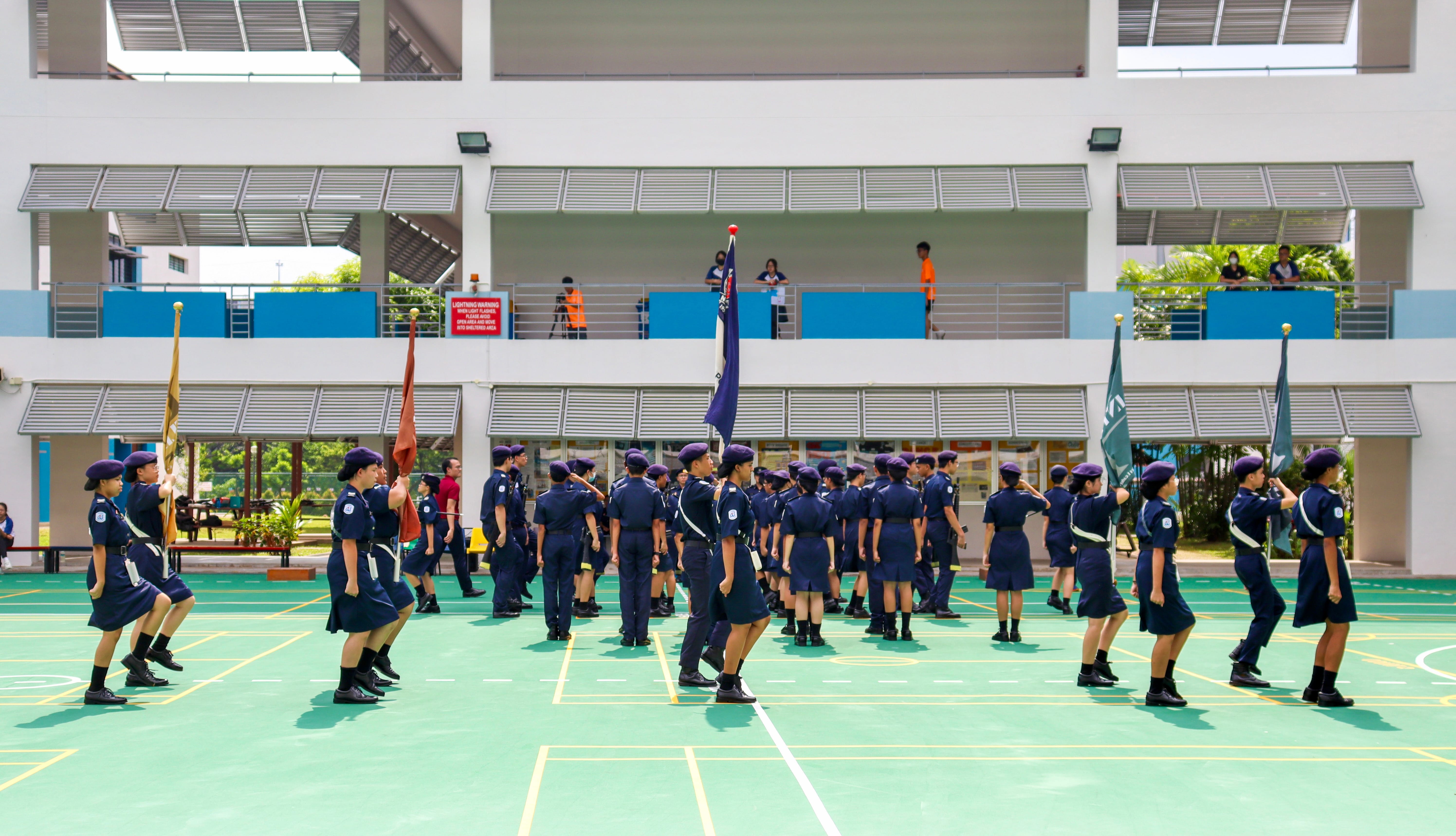 Uniformed cadets march with flags on a court in front of a school building with balconies and onlookers.