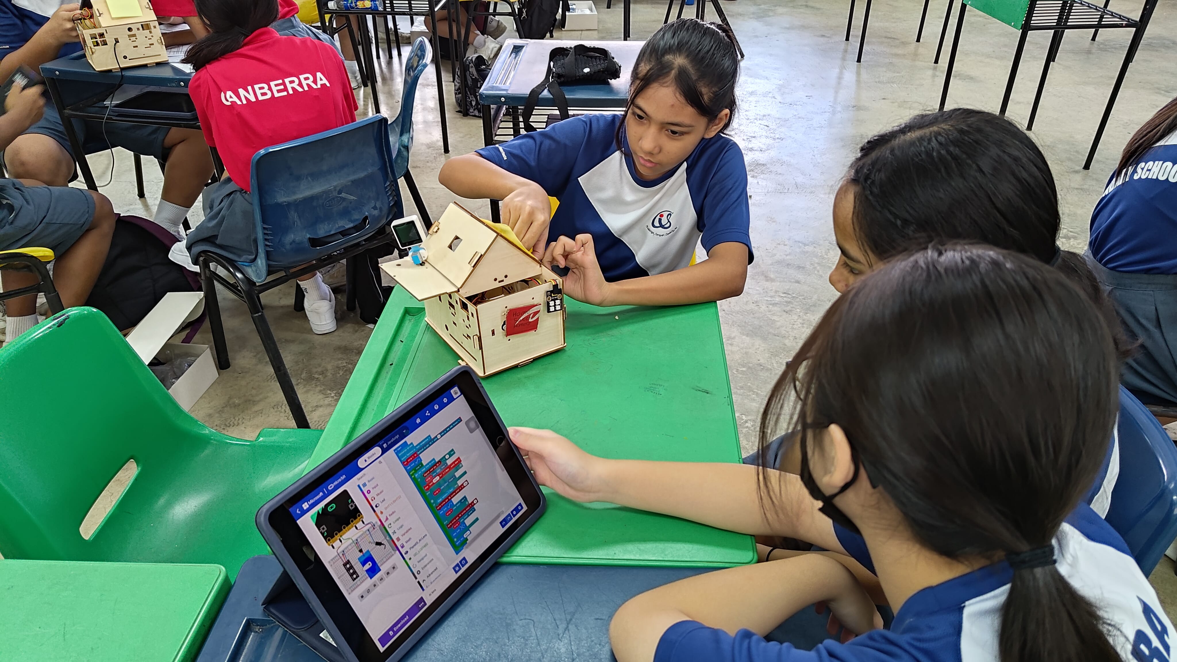 Students in school uniforms work on a wooden model house and a tablet with block coding software at a green desk.