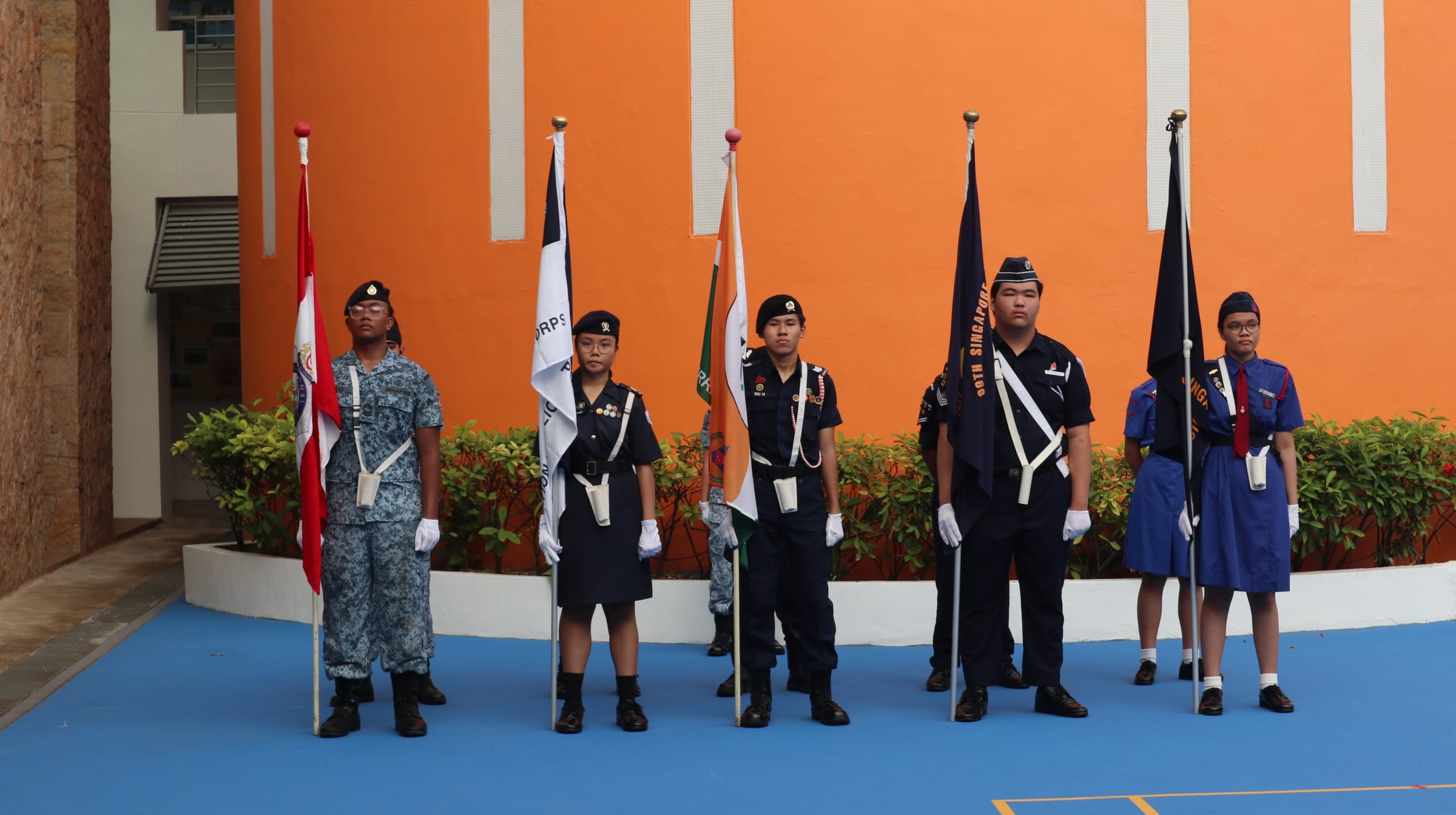 Six uniformed youth cadets stand at attention holding flags in front of an orange wall on a blue floor.