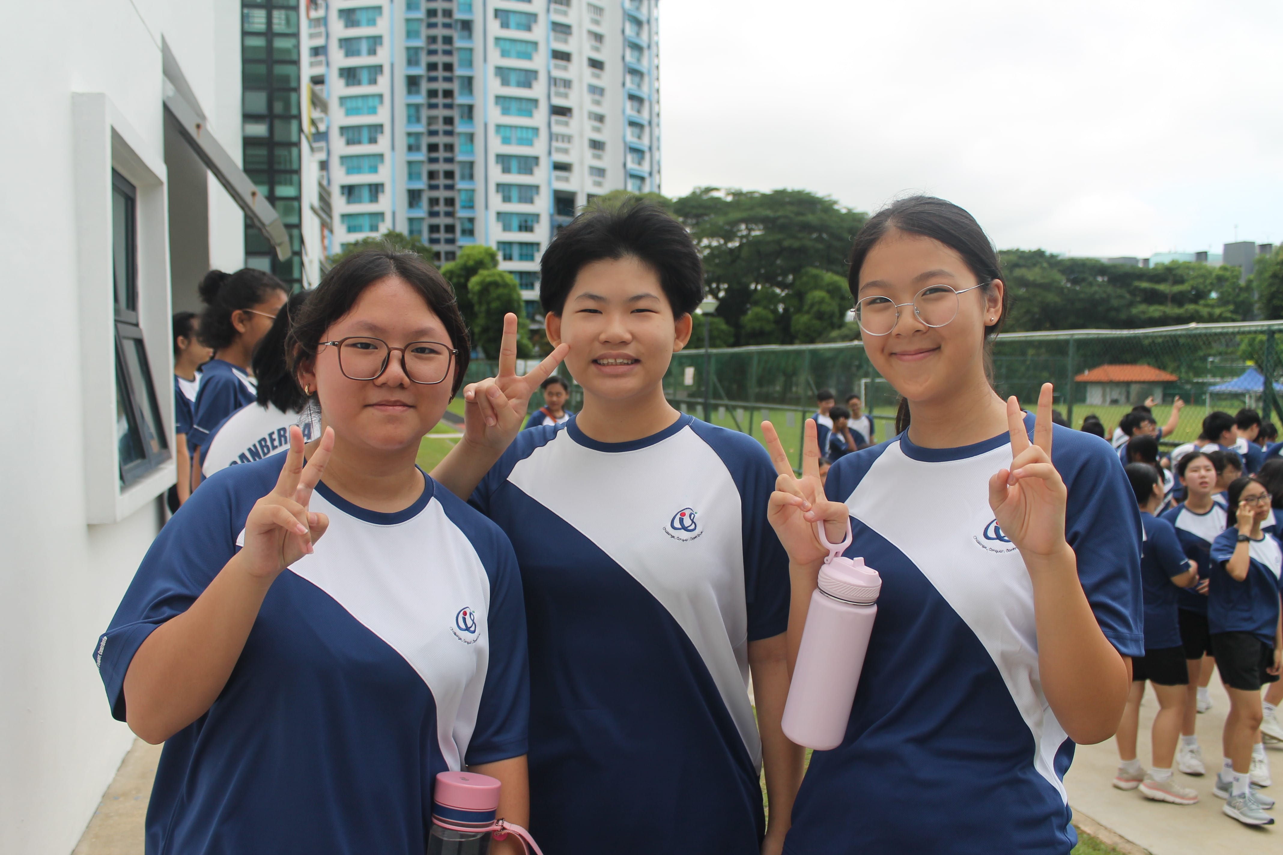 Three smiling students in Canberra Secondary School sports attire pose for a photo outdoors, each making a peace sign with their fingers. Two of them are holding water bottles. Other students and a school building are visible in the background.