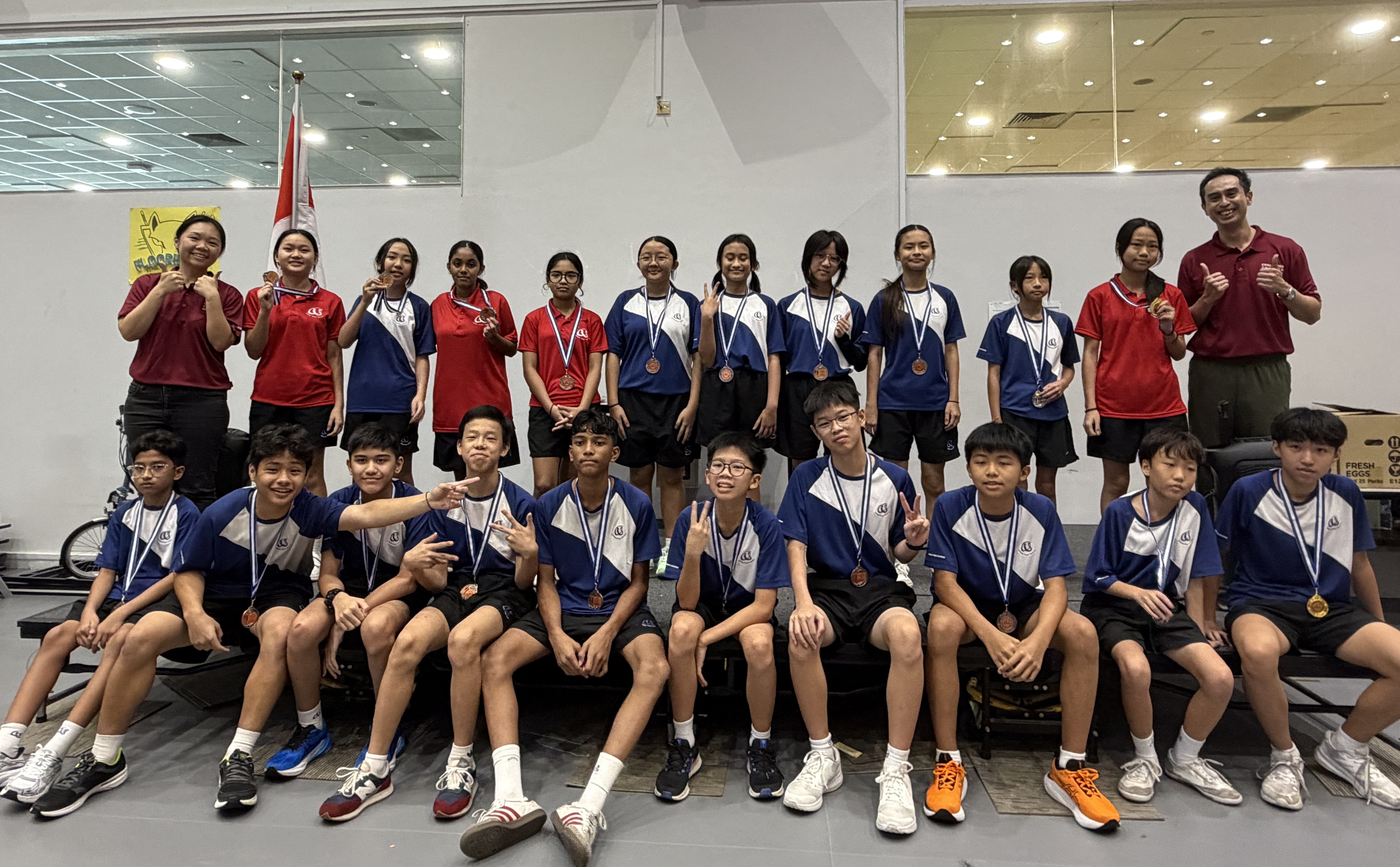 A large group of secondary school students in blue-and-white and red sports uniforms pose for a group photo, many displaying medals around their necks. Two teachers in maroon polo shirts flank the group.