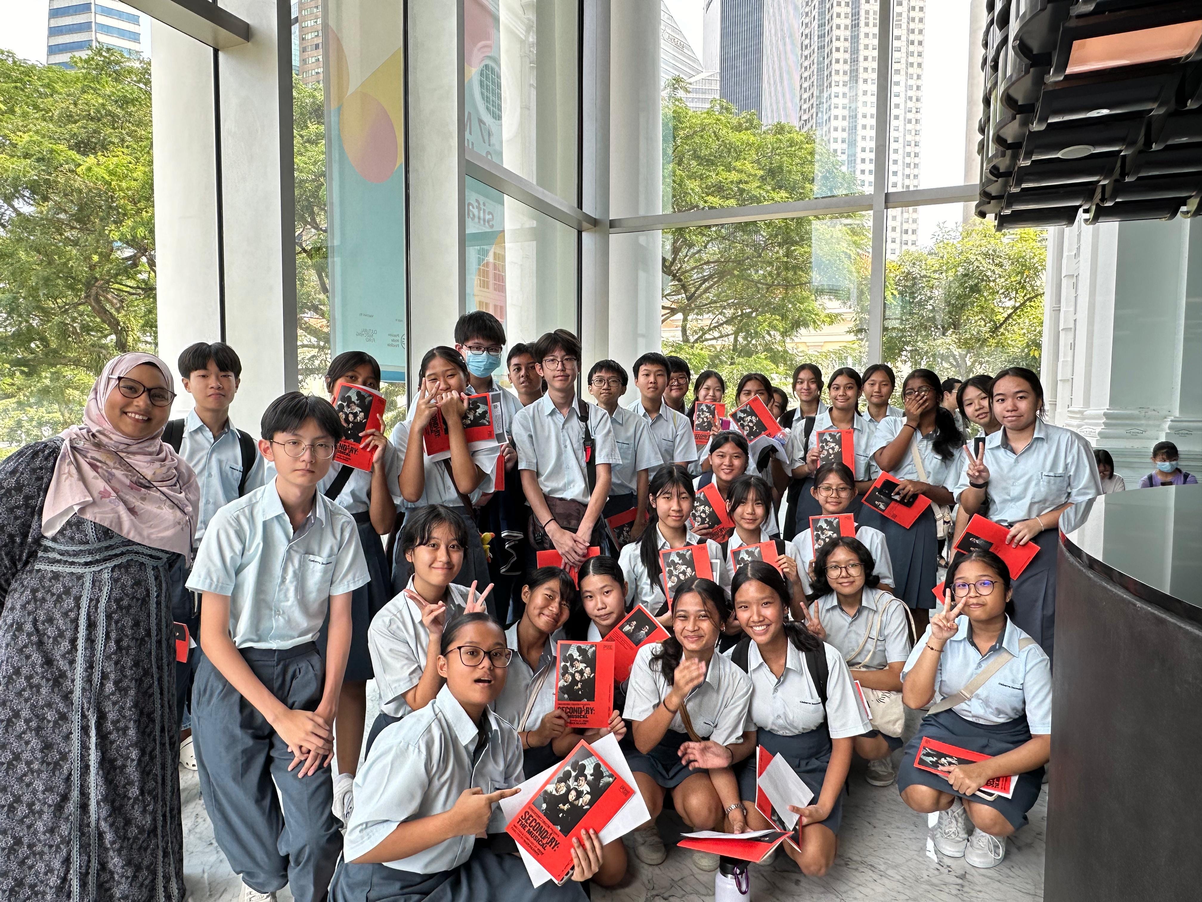 A large group of students in light blue uniforms holding red pamphlets pose together indoors near tall windows.