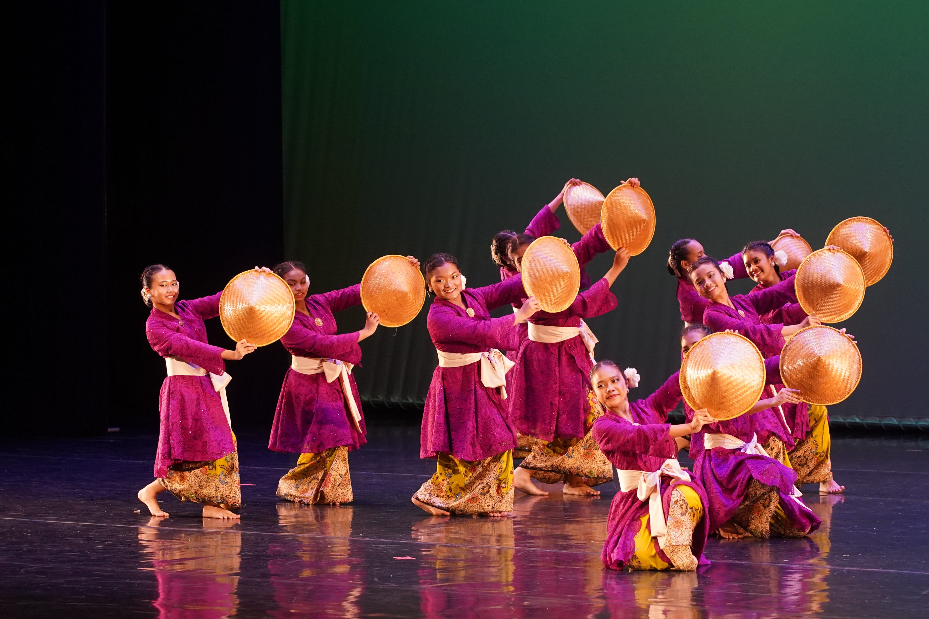 Girls in purple traditional costumes hold woven conical hats while performing a cultural dance on a lit stage.