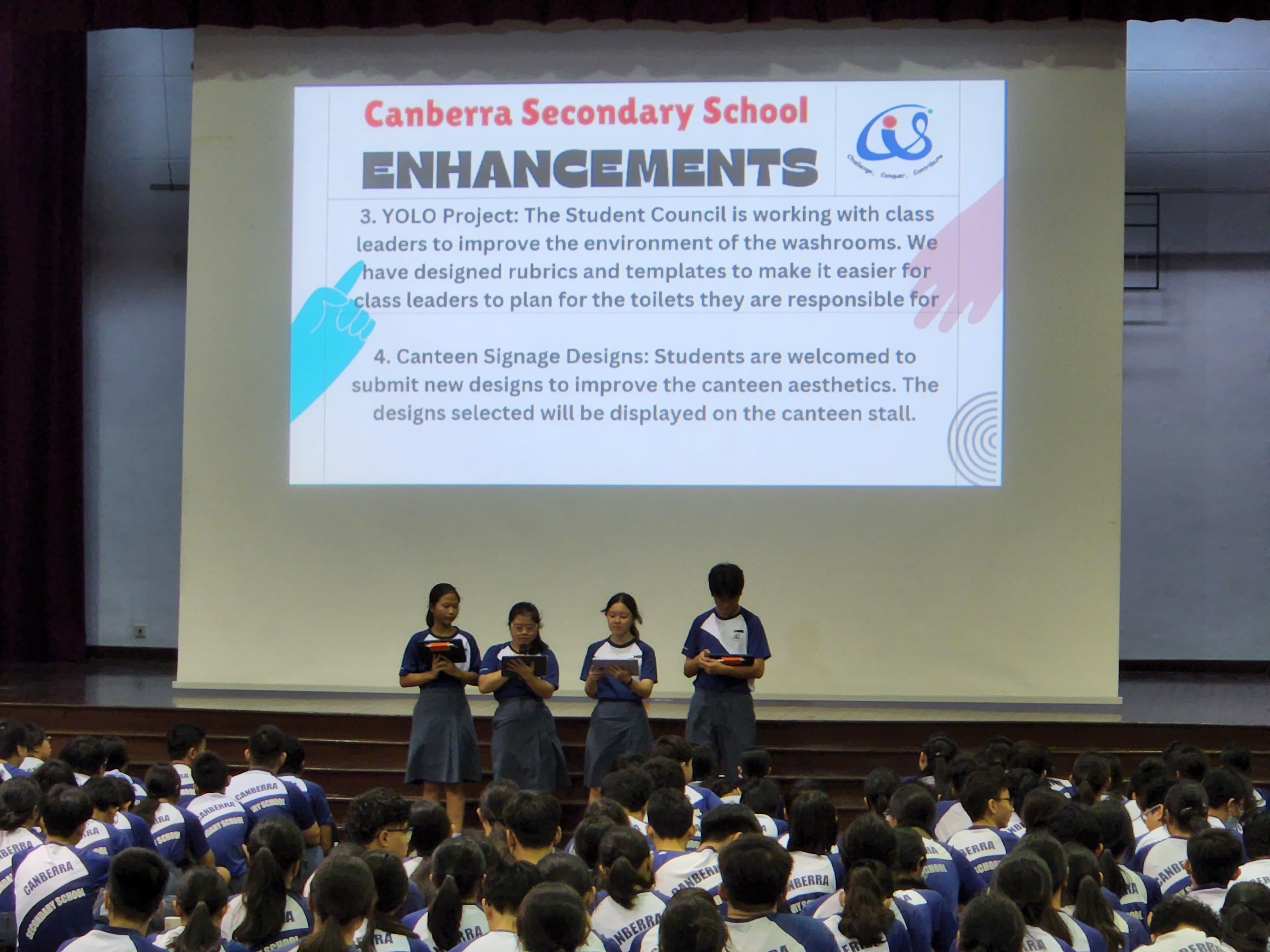 Four students address the school in the school hall as a large screen has talking points on school enhacements