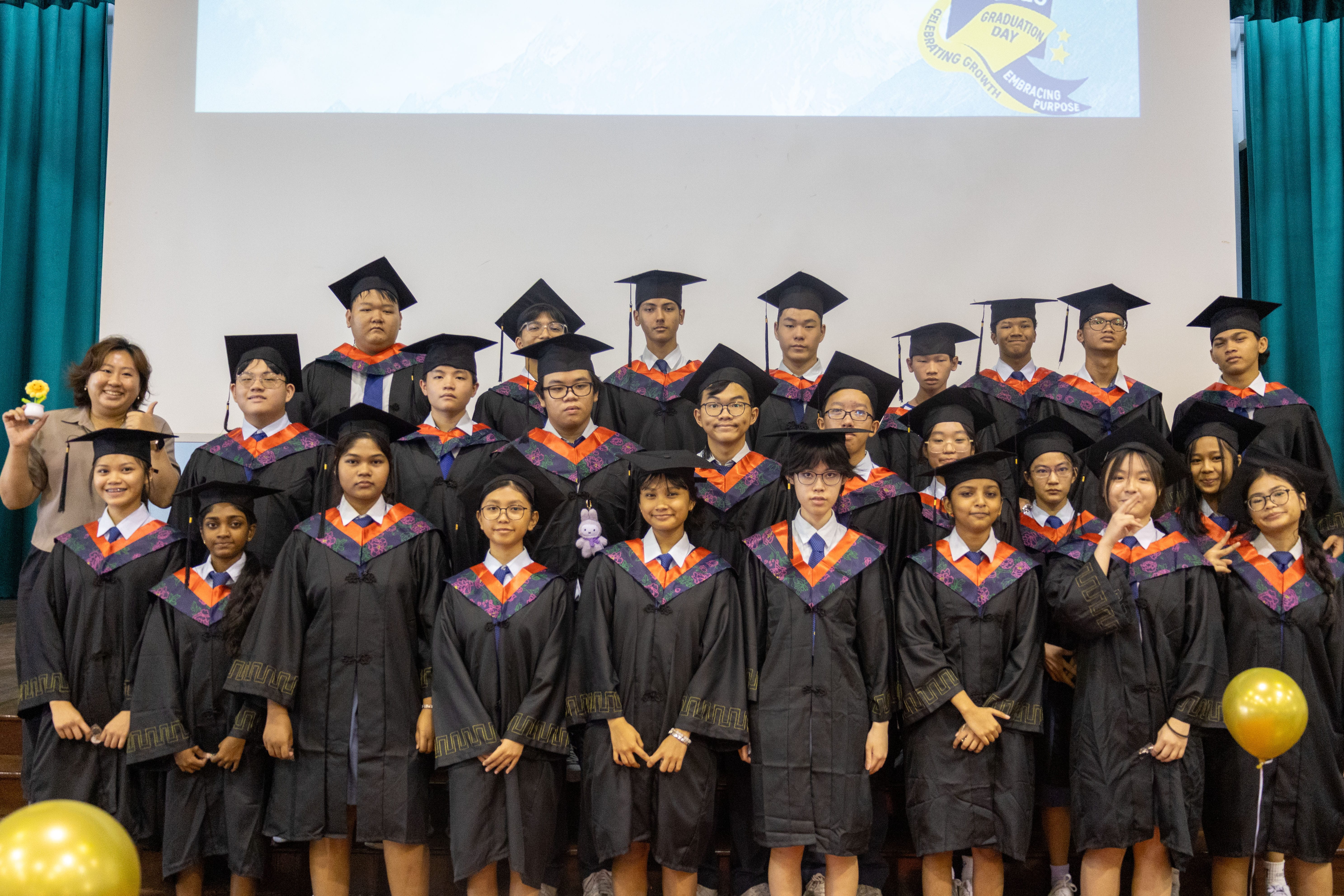 Group of graduates in black caps and gowns posing on stage; teacher at left; yellow balloons in front.