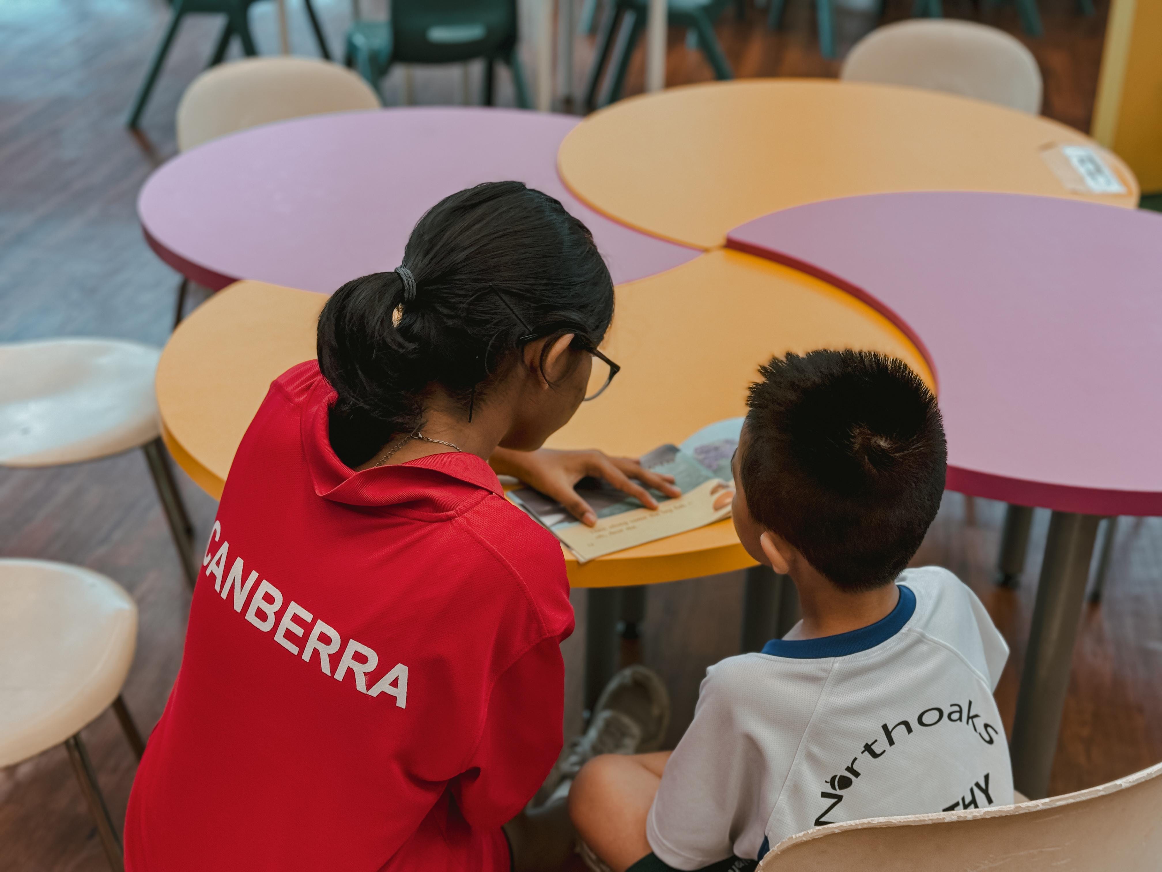 A secondary school student in a red tee reads a picture book to a primary school student in a white tee
