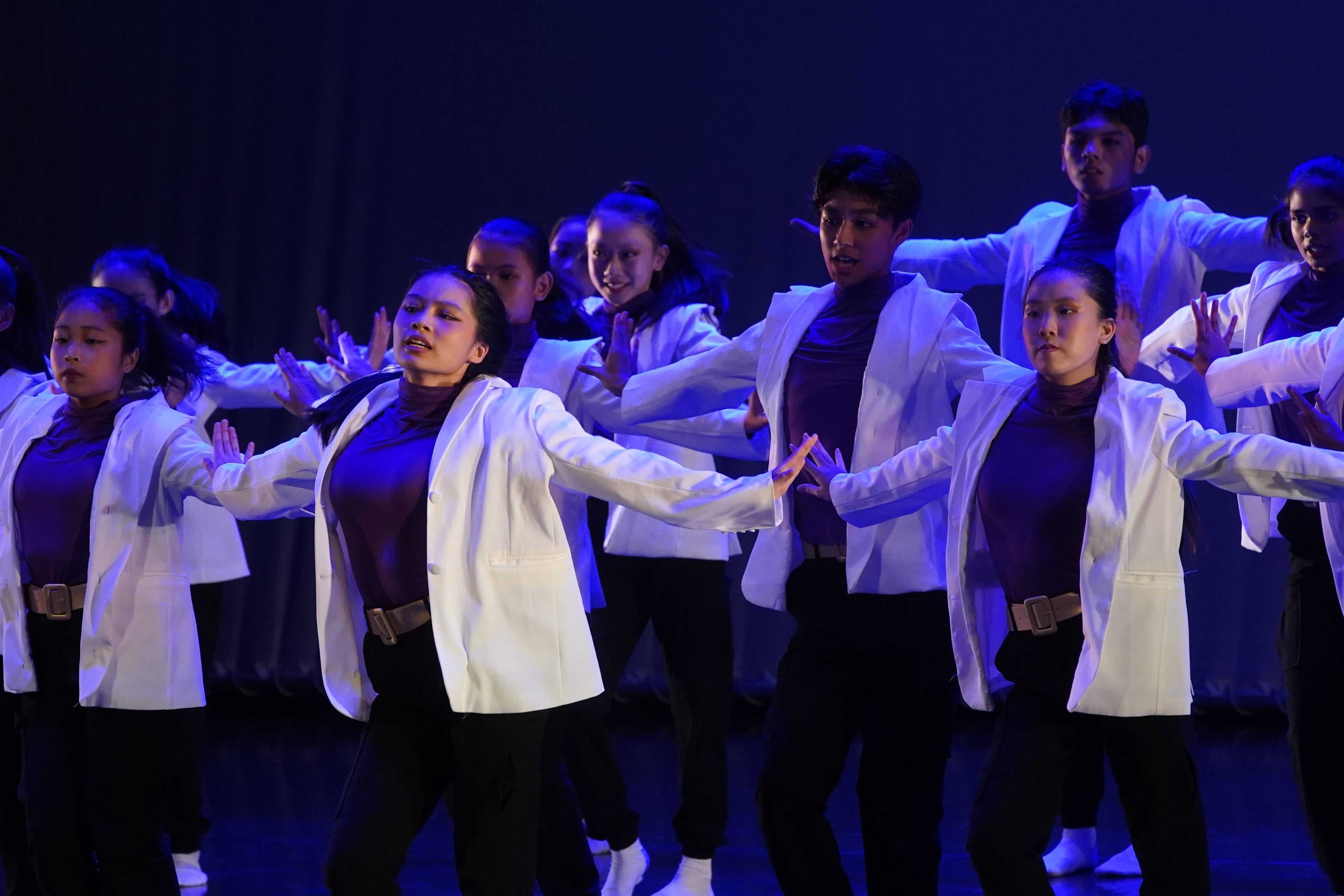 A large group of dancers in white blazers and purple tops perform in unison on a dimly lit stage.