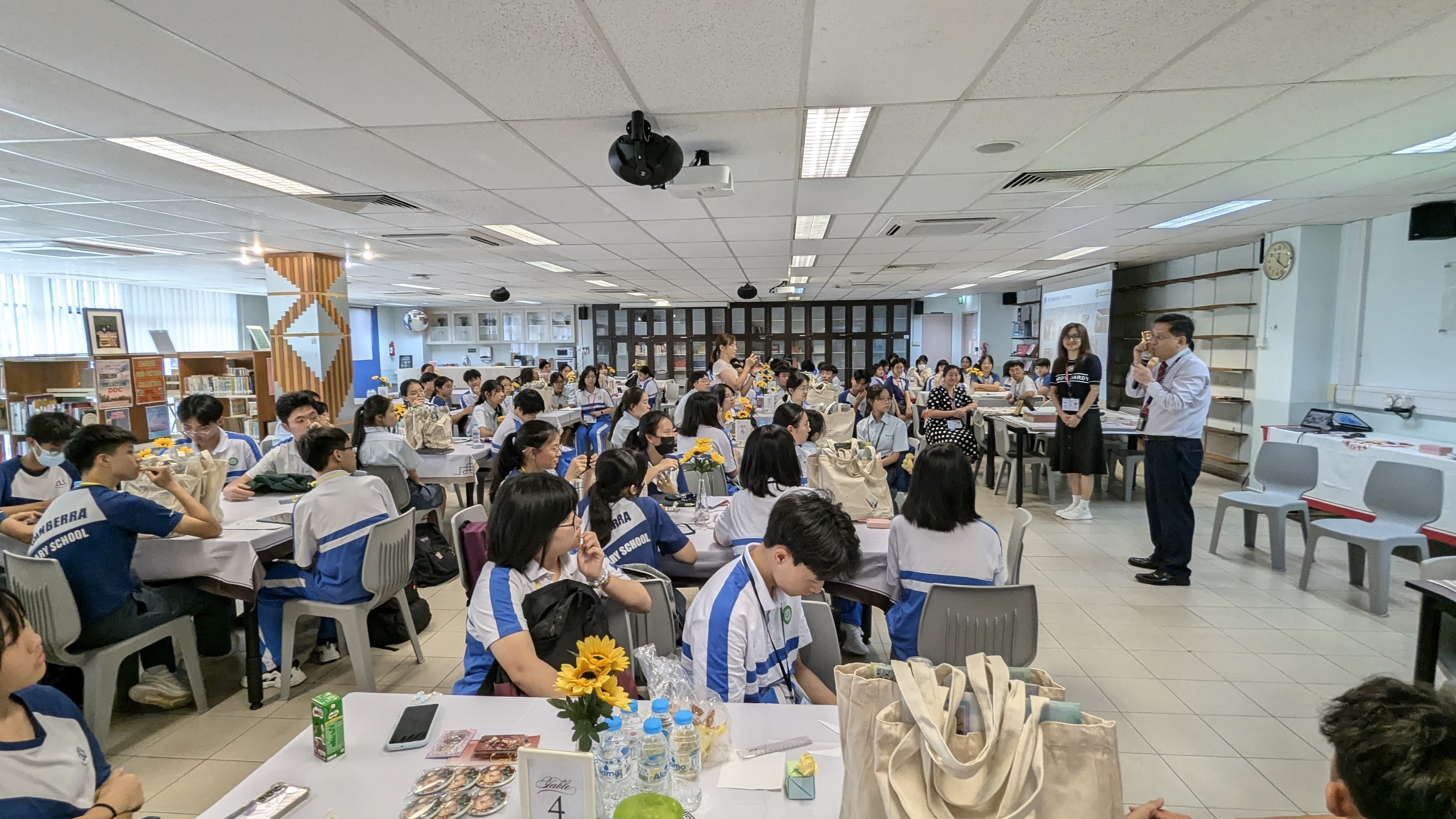 Students in blue and white uniforms sit at tables in a school hall while two adults stand and present.