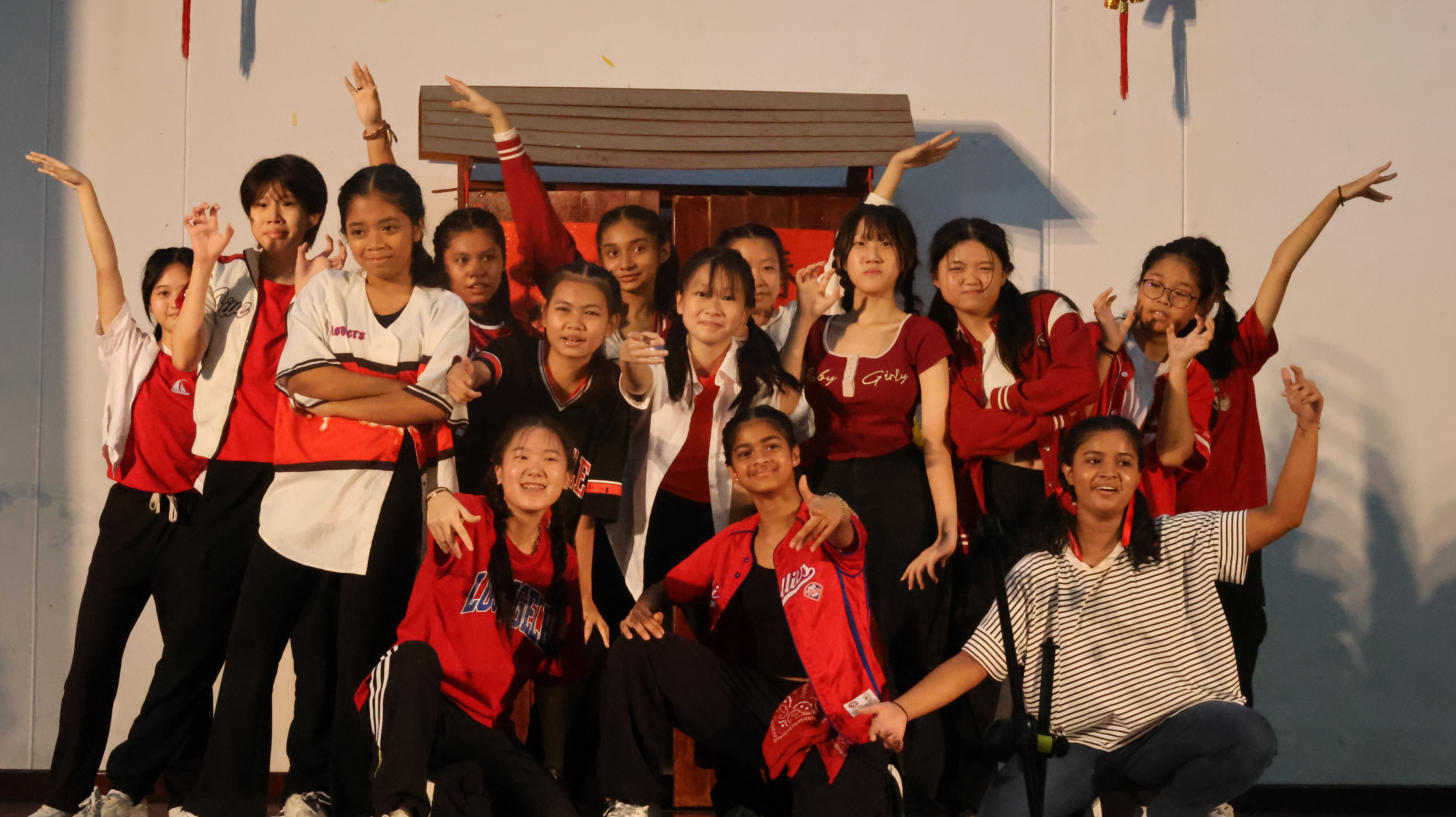 A group of teenage girls in red and white outfits pose energetically on a stage, some raising their arms.