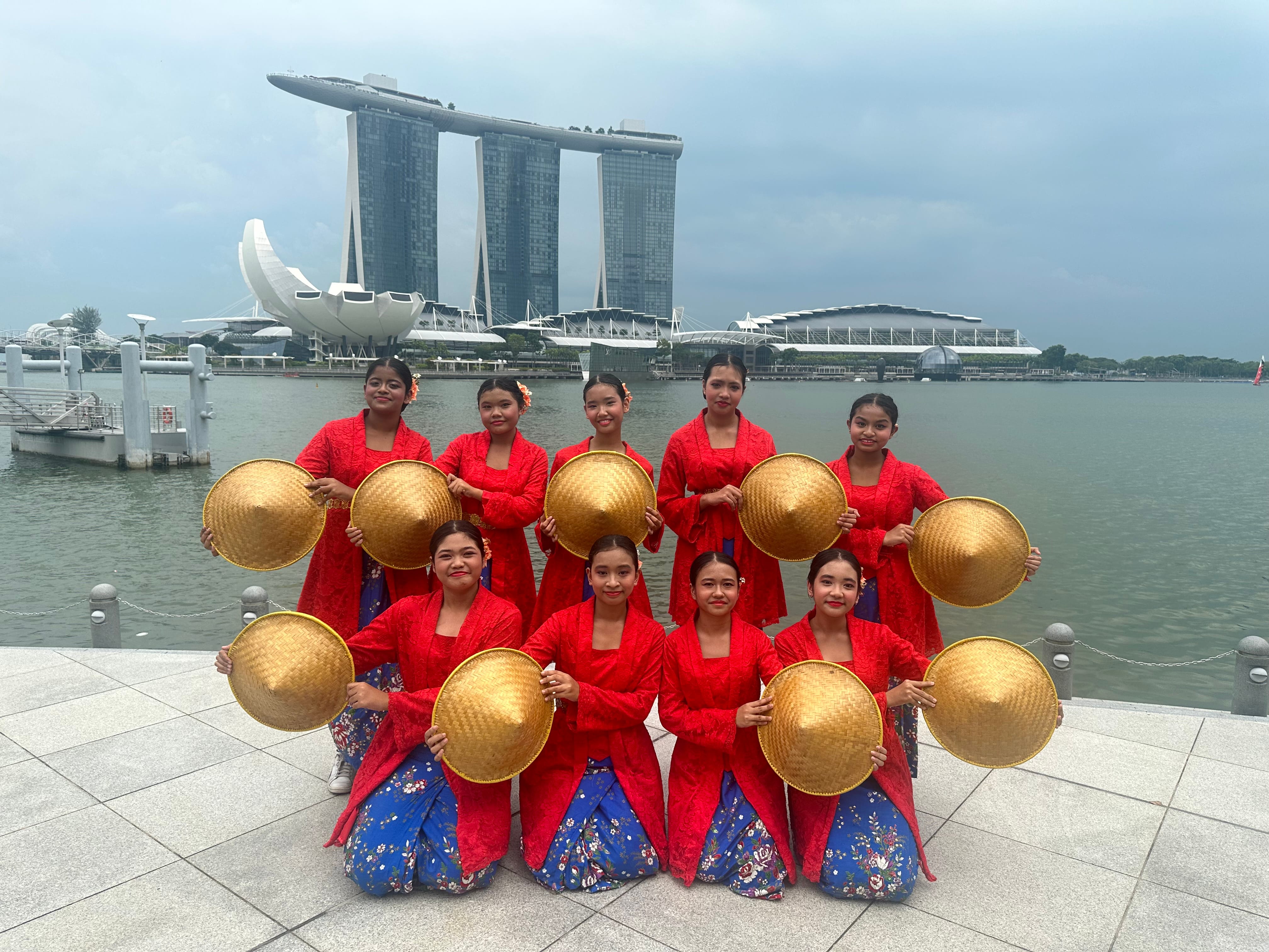Ten women in red outfits hold round straw hats by the water, with Marina Bay Sands in the background.