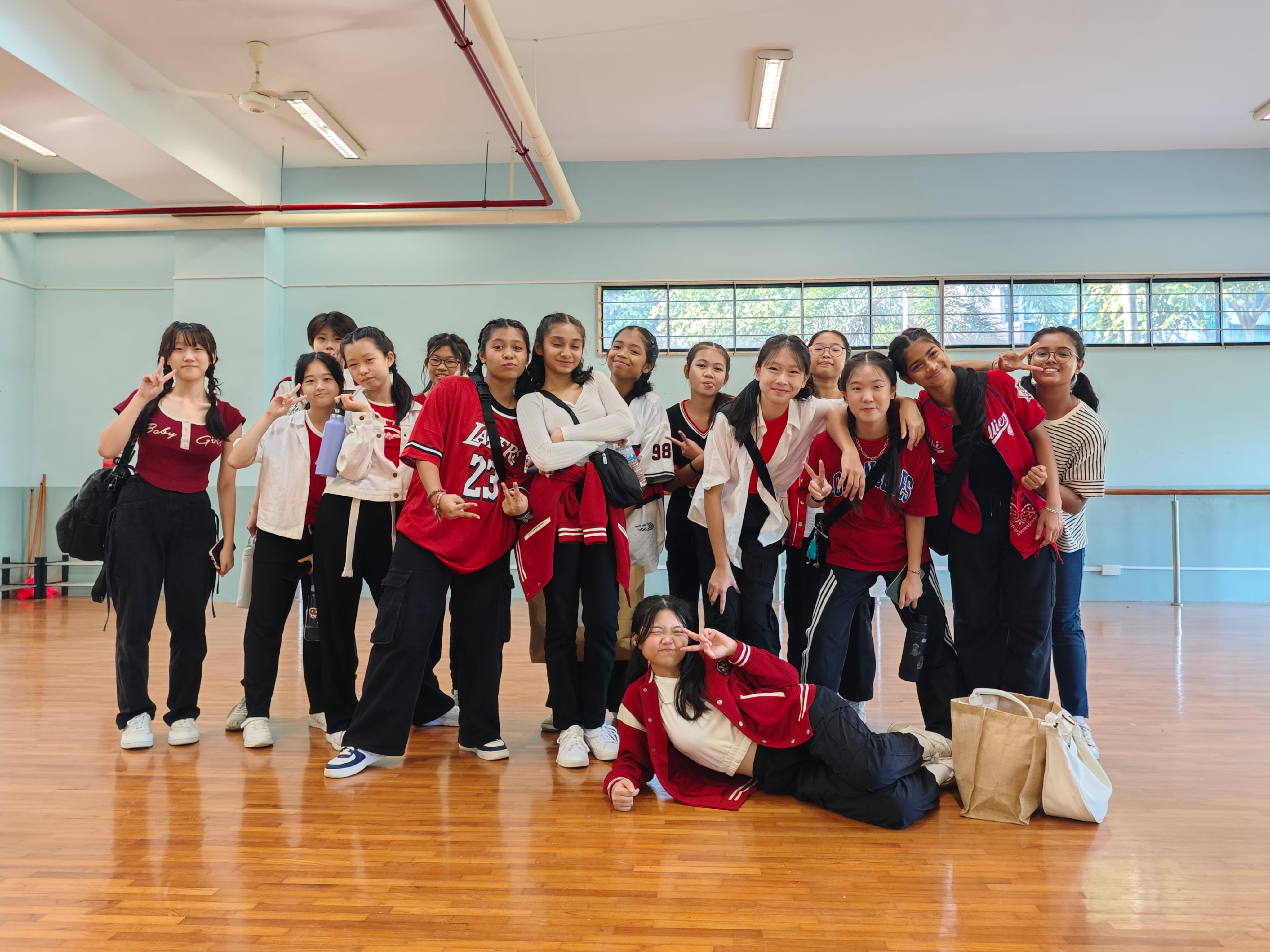A group of teenage girls in red and black outfits pose with peace signs in a dance studio with wooden floors.