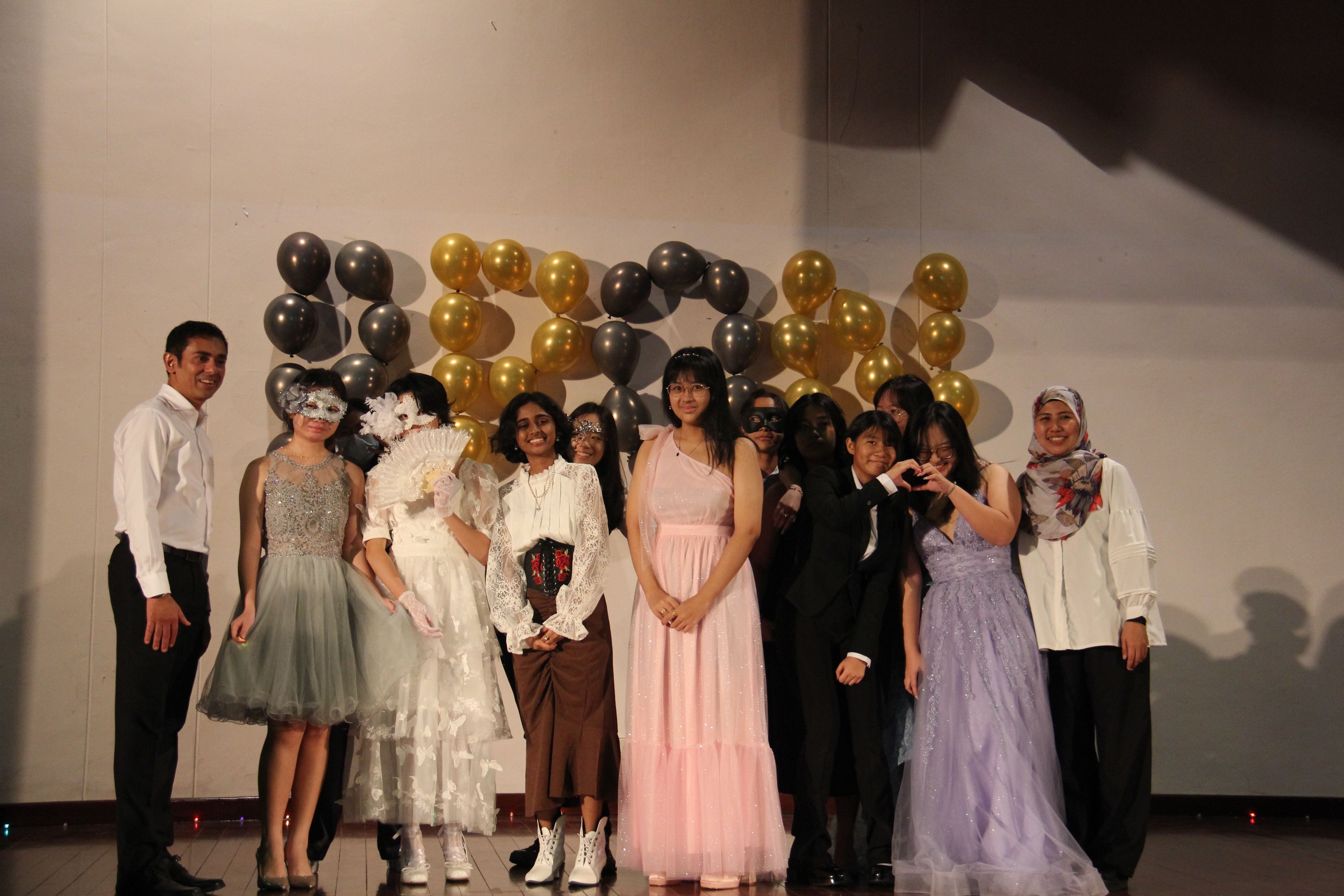 A group of about twelve dressed-up teens pose in front of gold and silver balloons at a formal event.