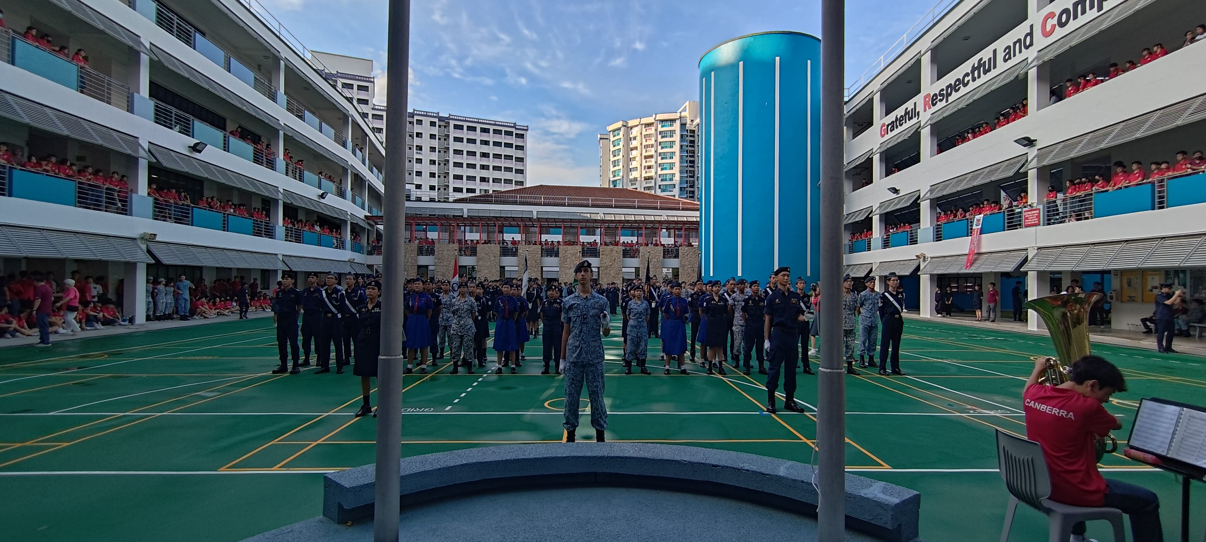 Uniformed students stand at attention in a school courtyard during a formal parade, watched by crowds on surrounding building balconies.