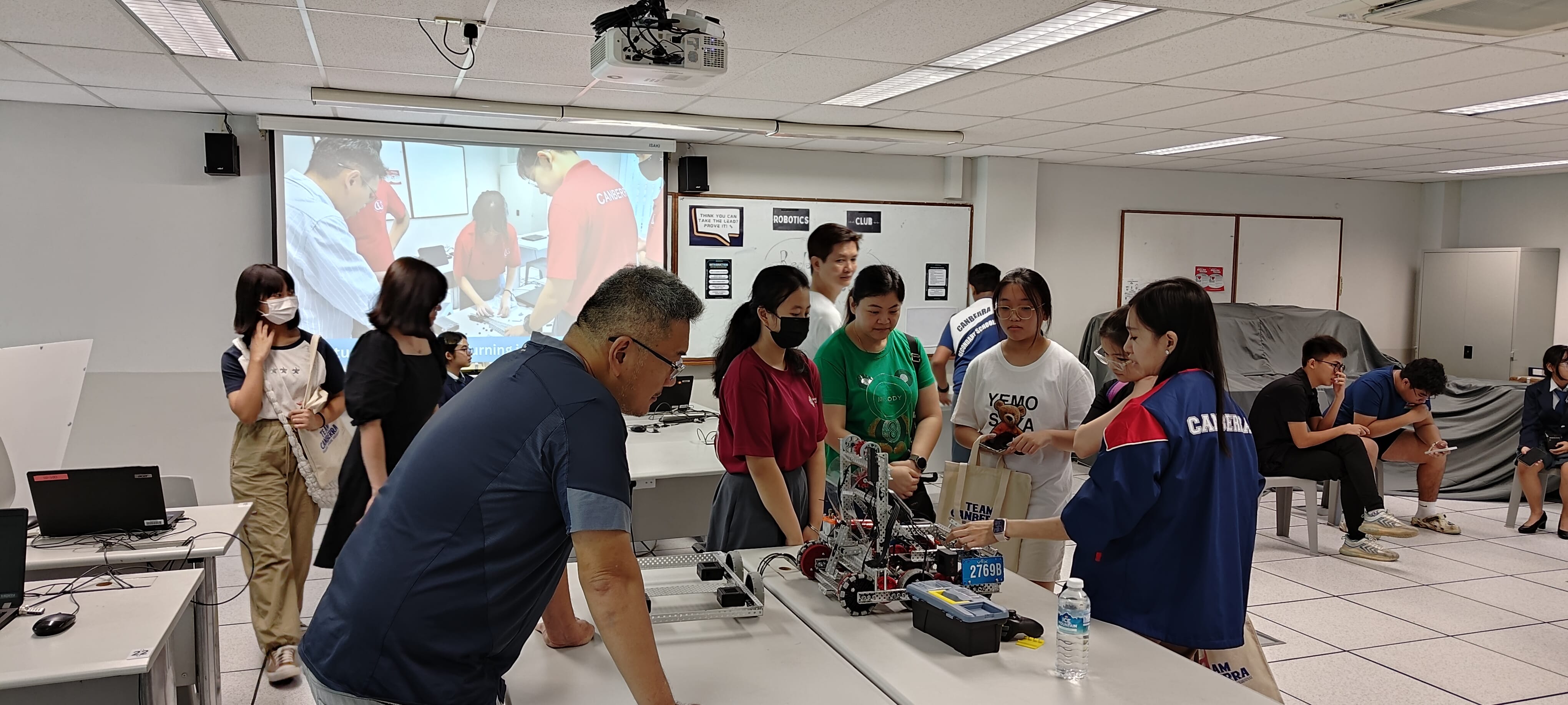 Students and an adult gather around a table examining a competition robot labeled 2769B in a robotics club classroom.