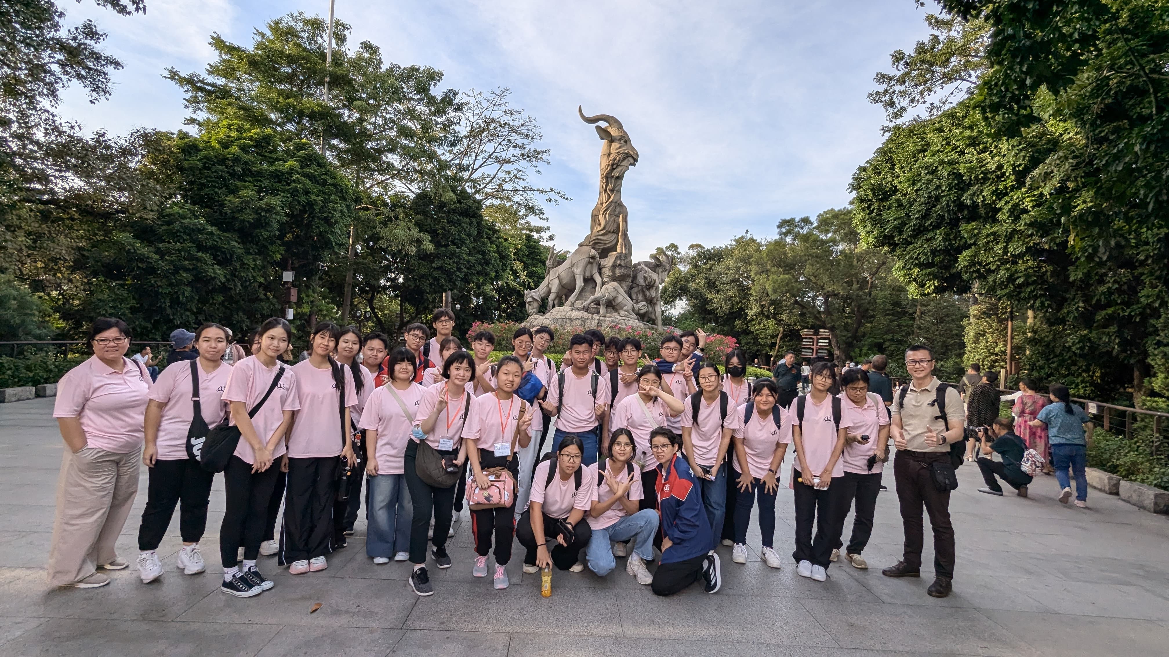 A large group in matching pink shirts poses in front of a tall bronze five-goat sculpture in a tree-lined park.