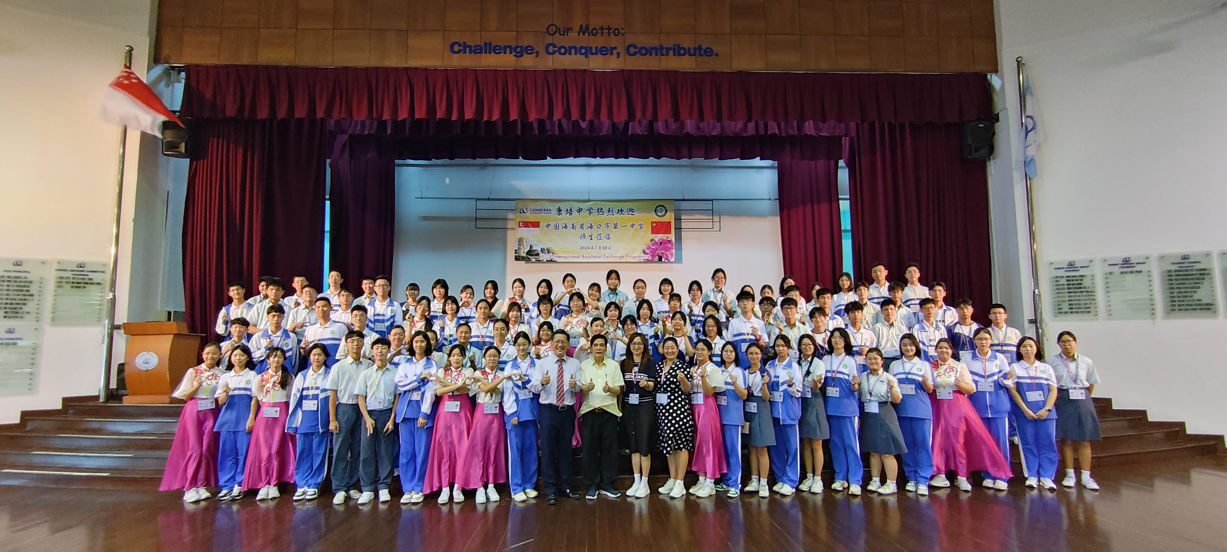 Large group of students and teachers posing in a school hall with a motto reading Challenge, Conquer, Contribute.