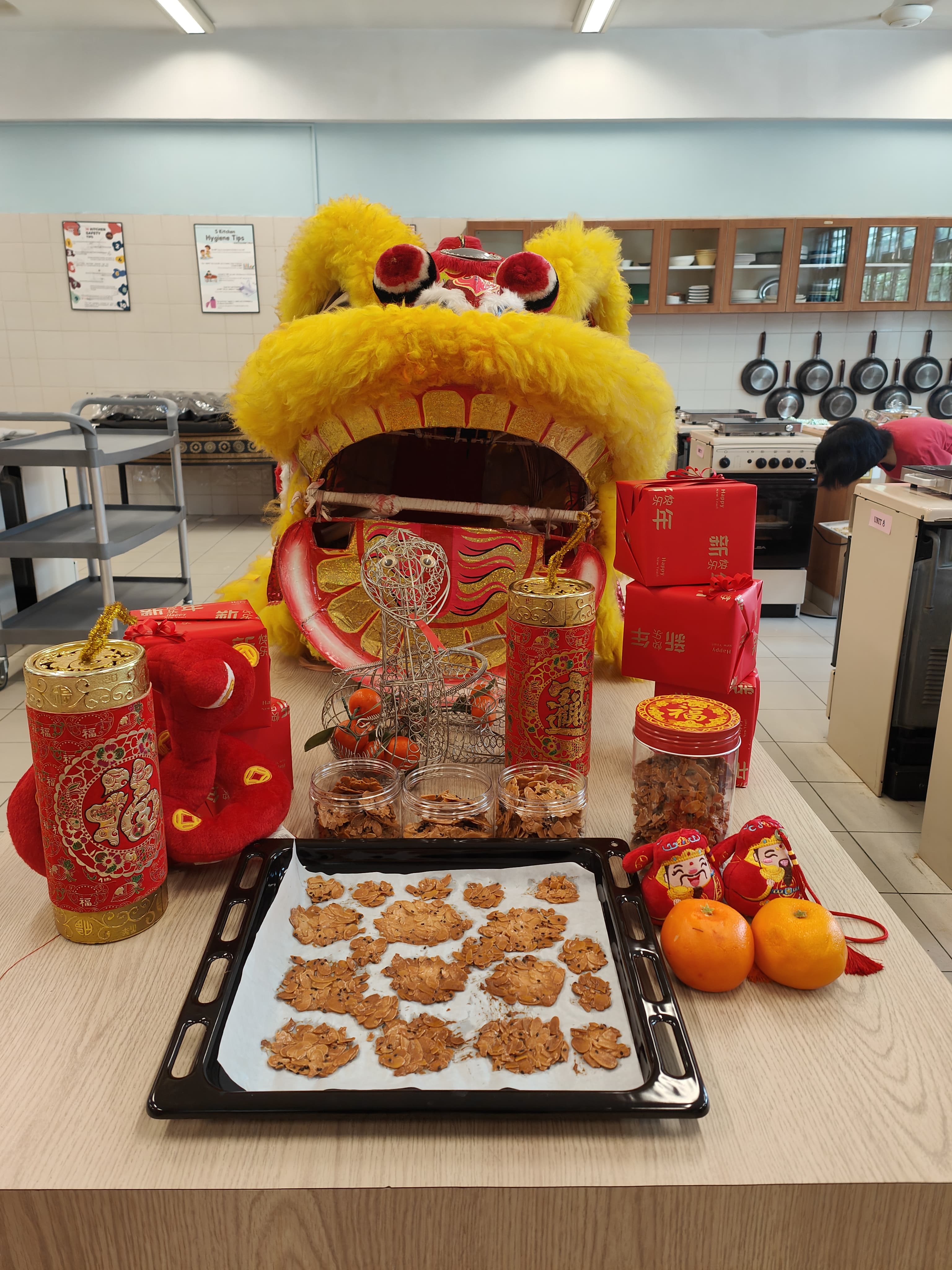 Yellow Chinese lion dance head on a table with cookies, oranges, red gift boxes, and festive decorations in a kitchen.