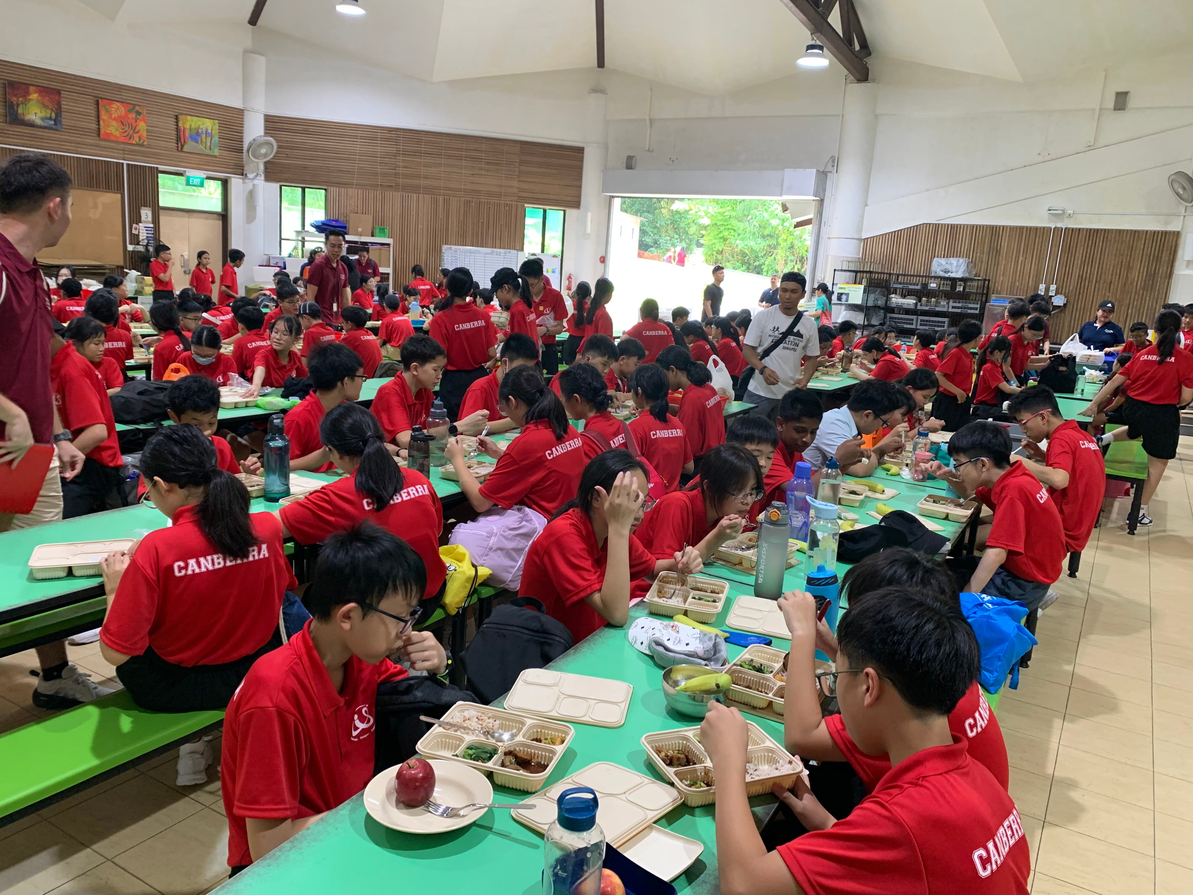 Students in red Canberra shirts eat meal trays at long green tables in a crowded school cafeteria.