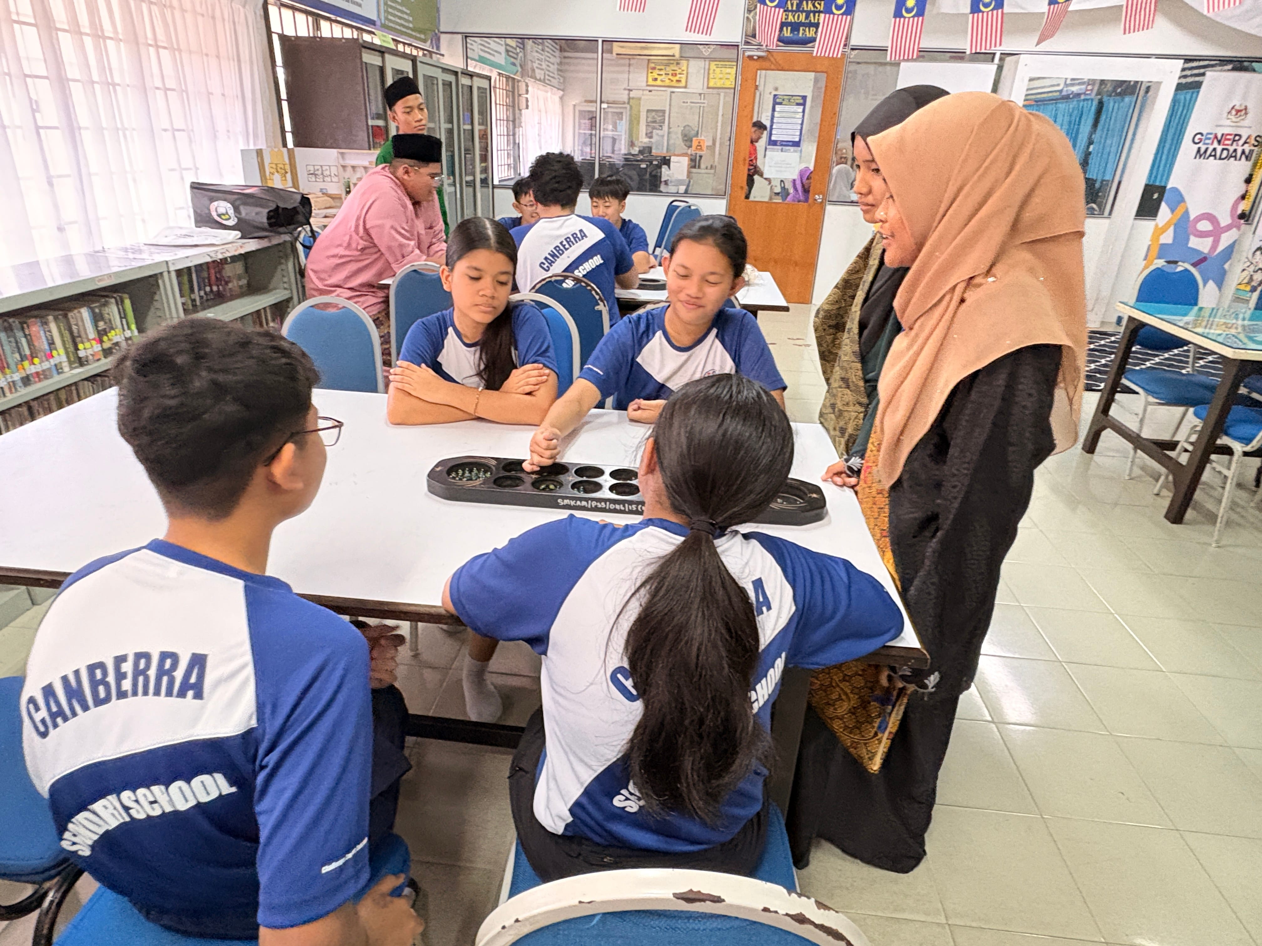 Students in Canberra school uniforms play a mancala board game at a table while two women observe.