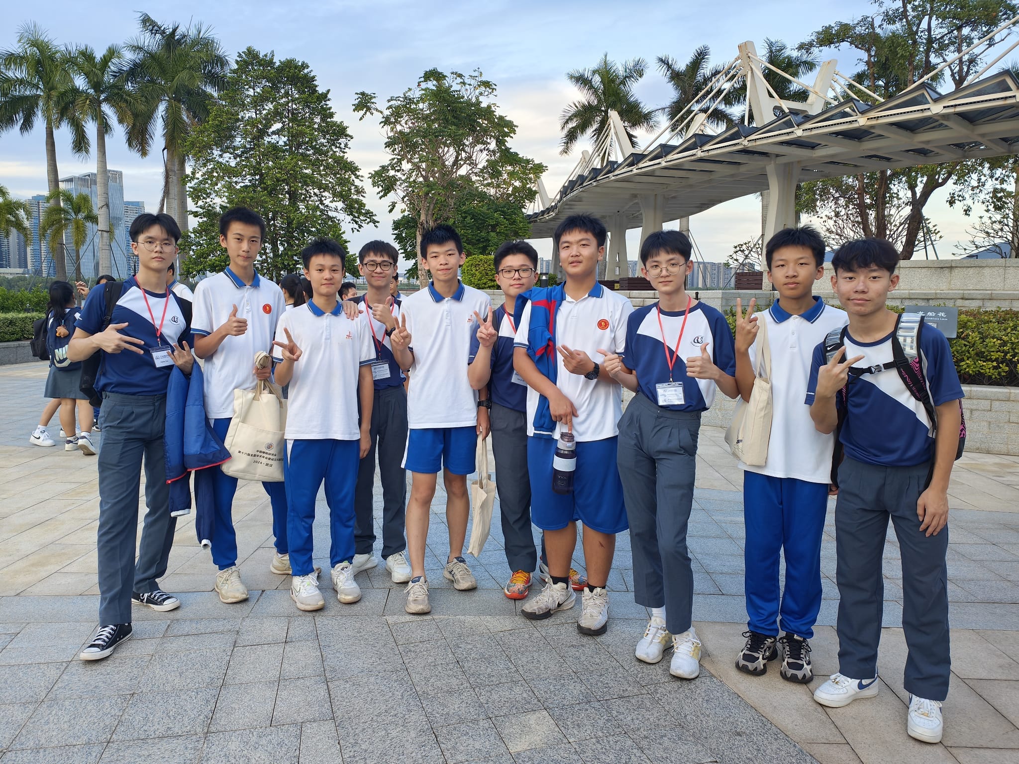 Nine students in blue and white uniforms pose outdoors making peace signs, with palm trees and a bridge behind them.
