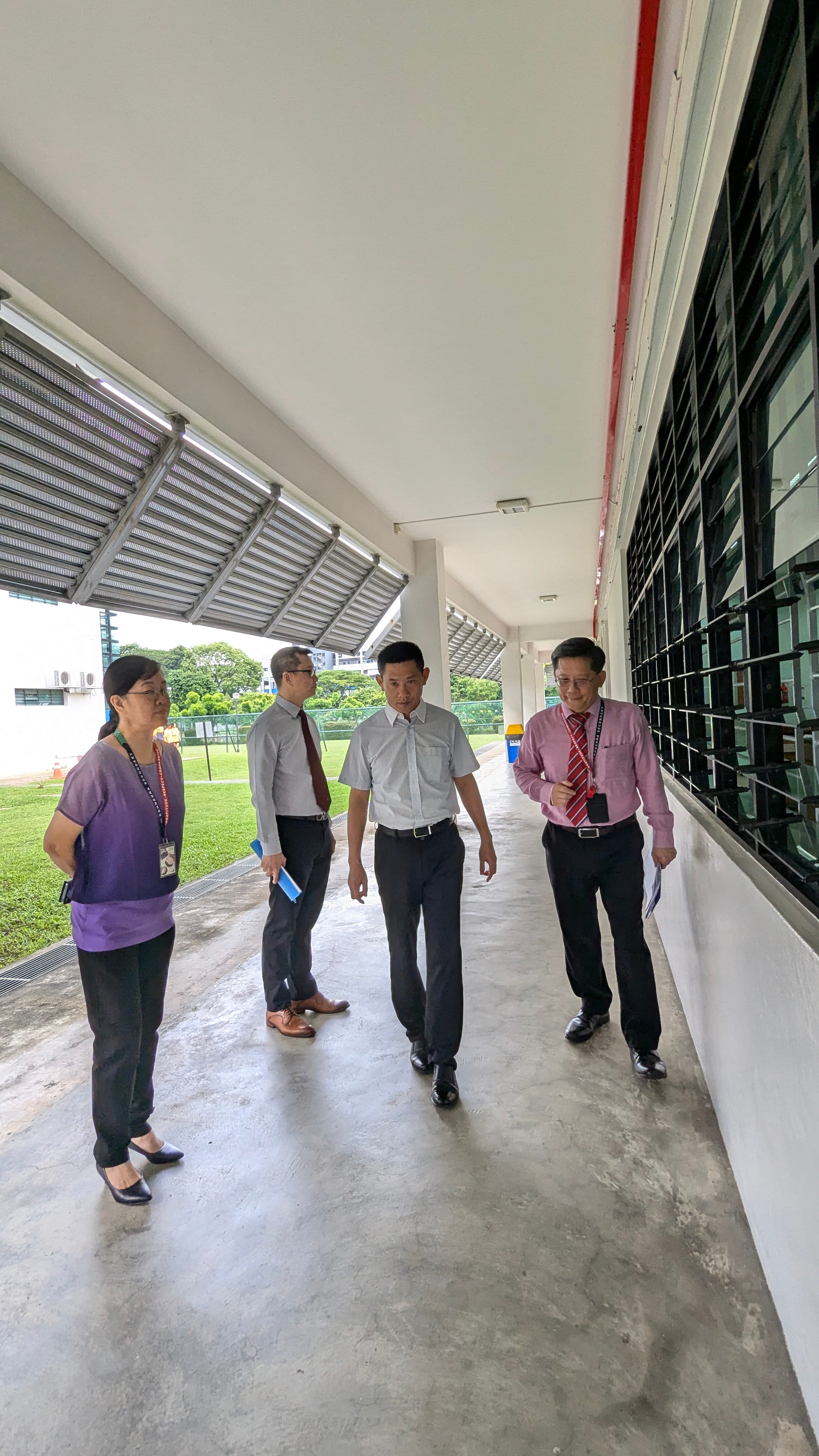 Four people walk along a covered corridor beside a building, with a green field visible in the background.