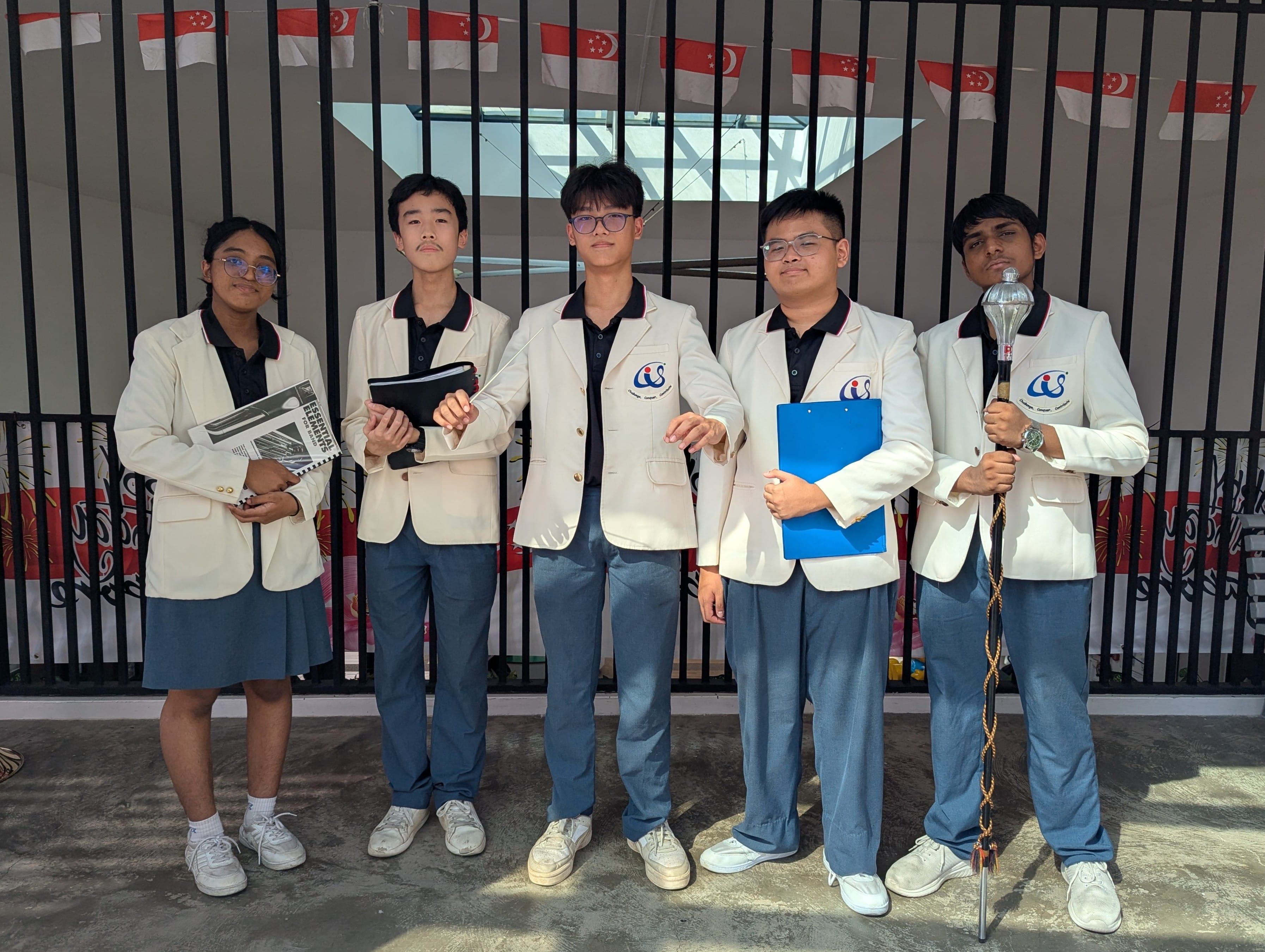 Five students in white blazers and blue uniforms pose together, with Singapore flags hanging in the background.