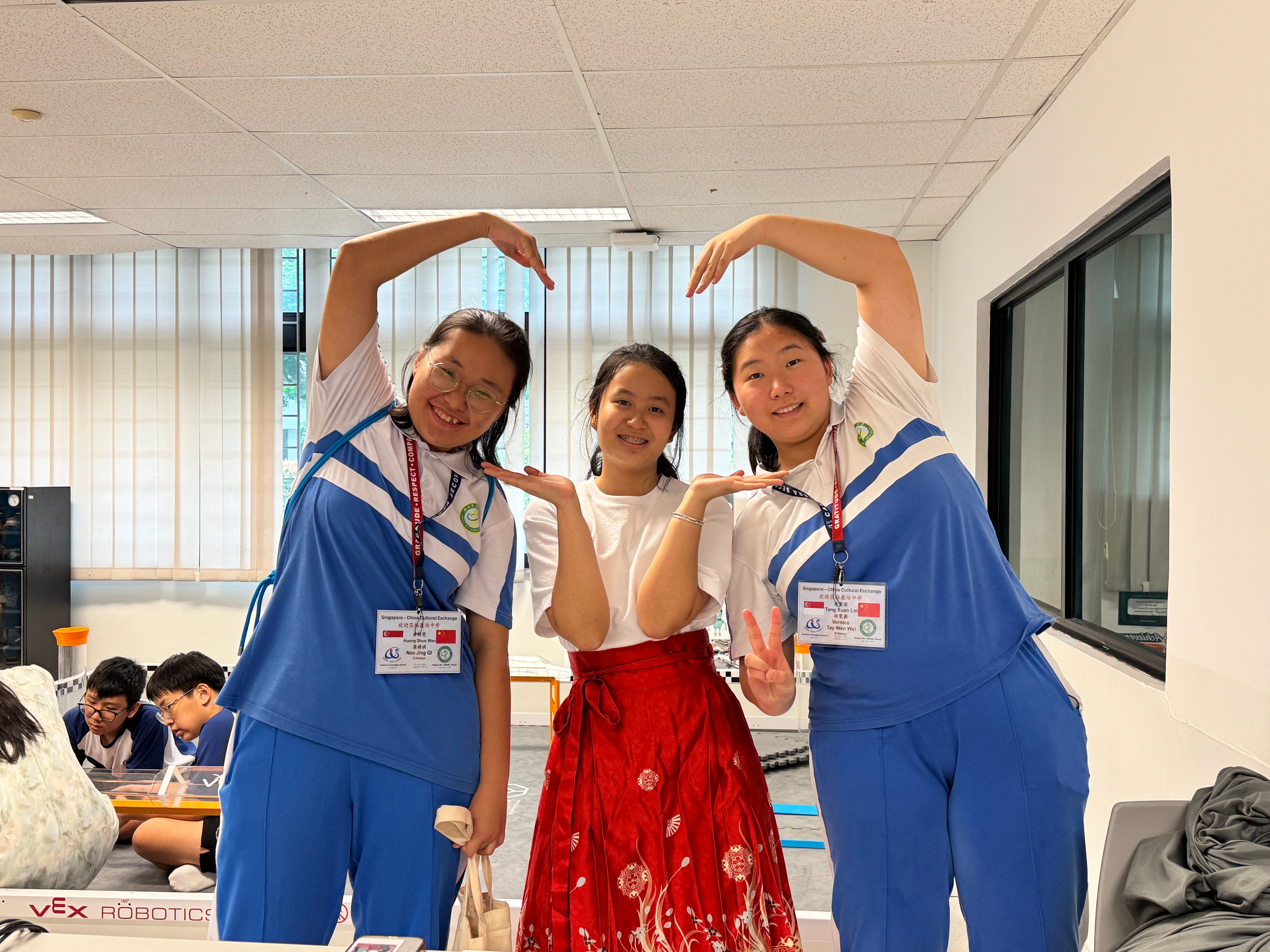 Three students pose indoors with arms forming hearts; two in blue uniforms, center in white top and red skirt.