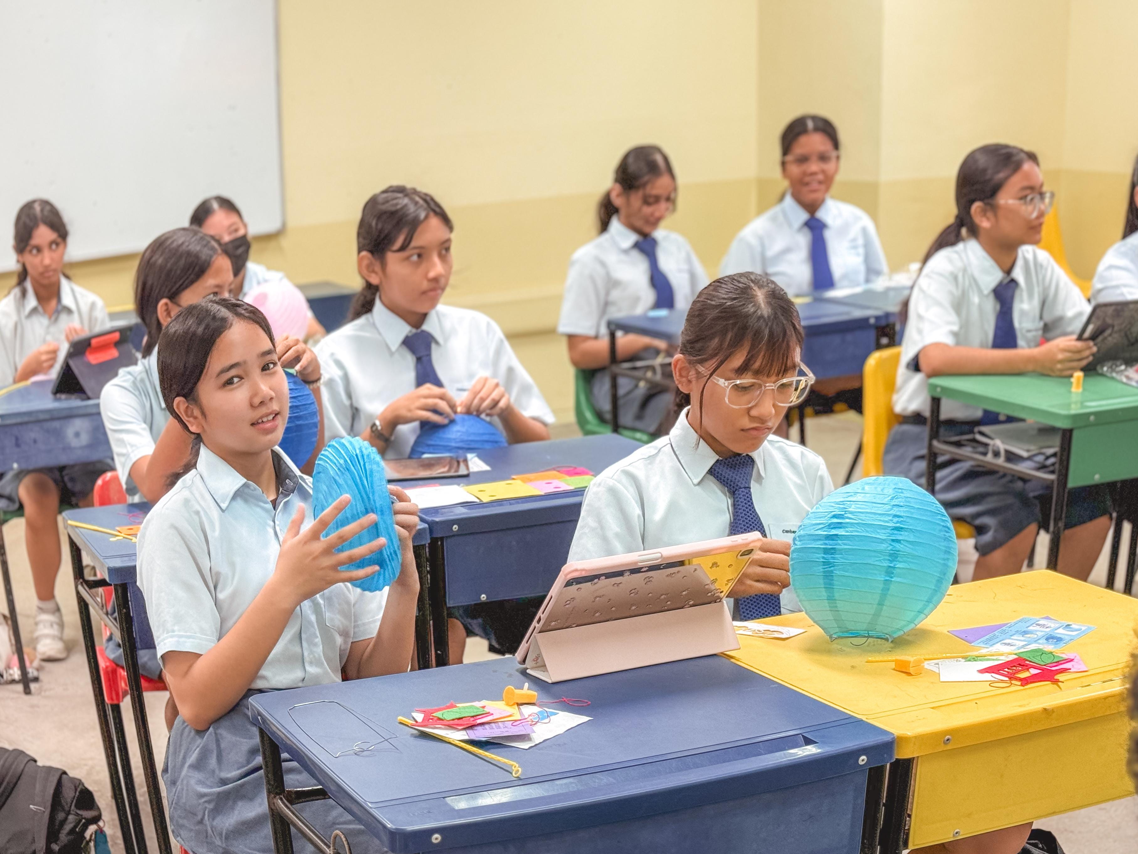 Students in school uniforms sit at colorful desks holding paper lanterns and tablets in a classroom.