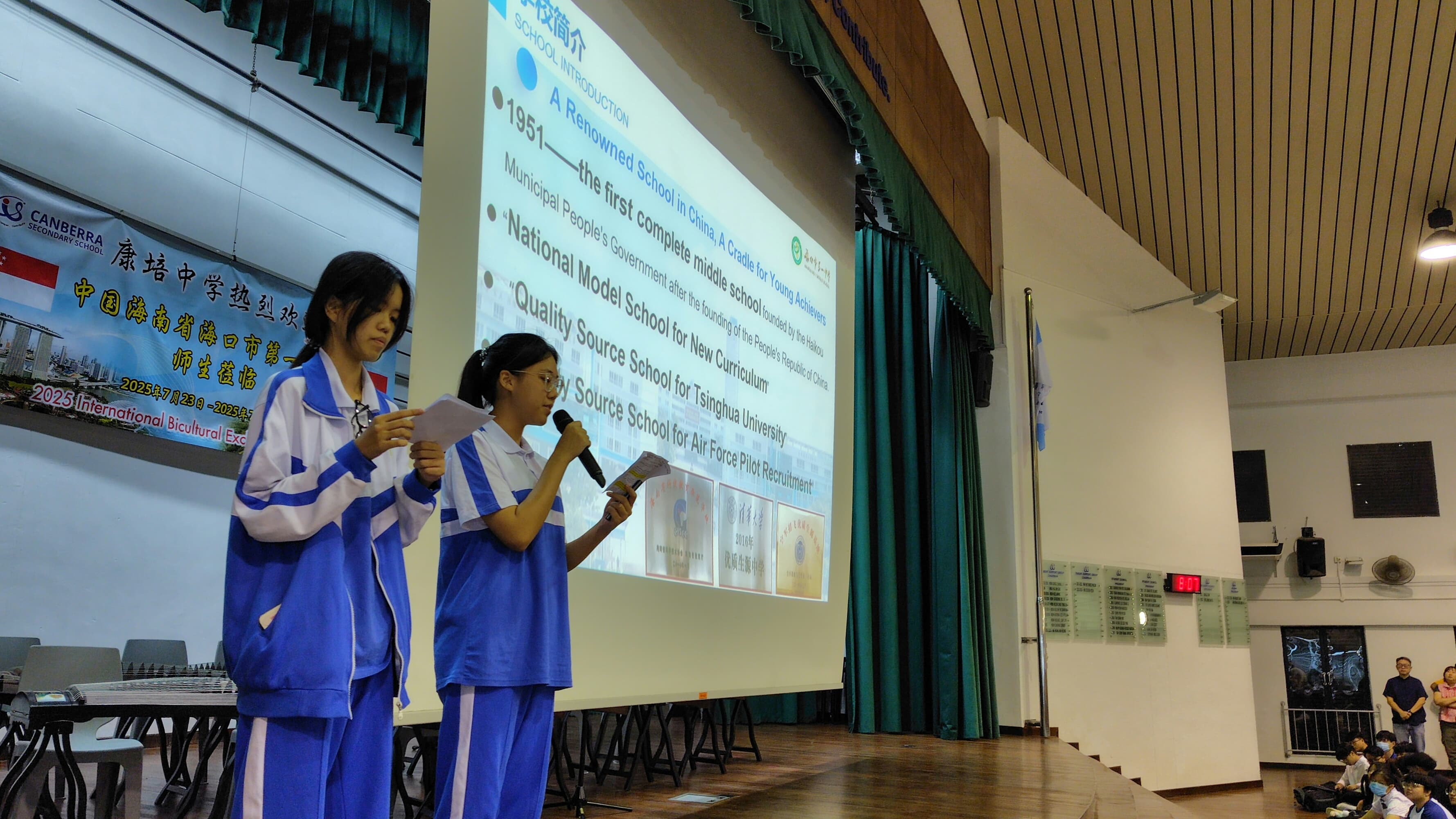 Two students in blue uniforms present on stage with a microphone, standing before a school introduction slideshow.