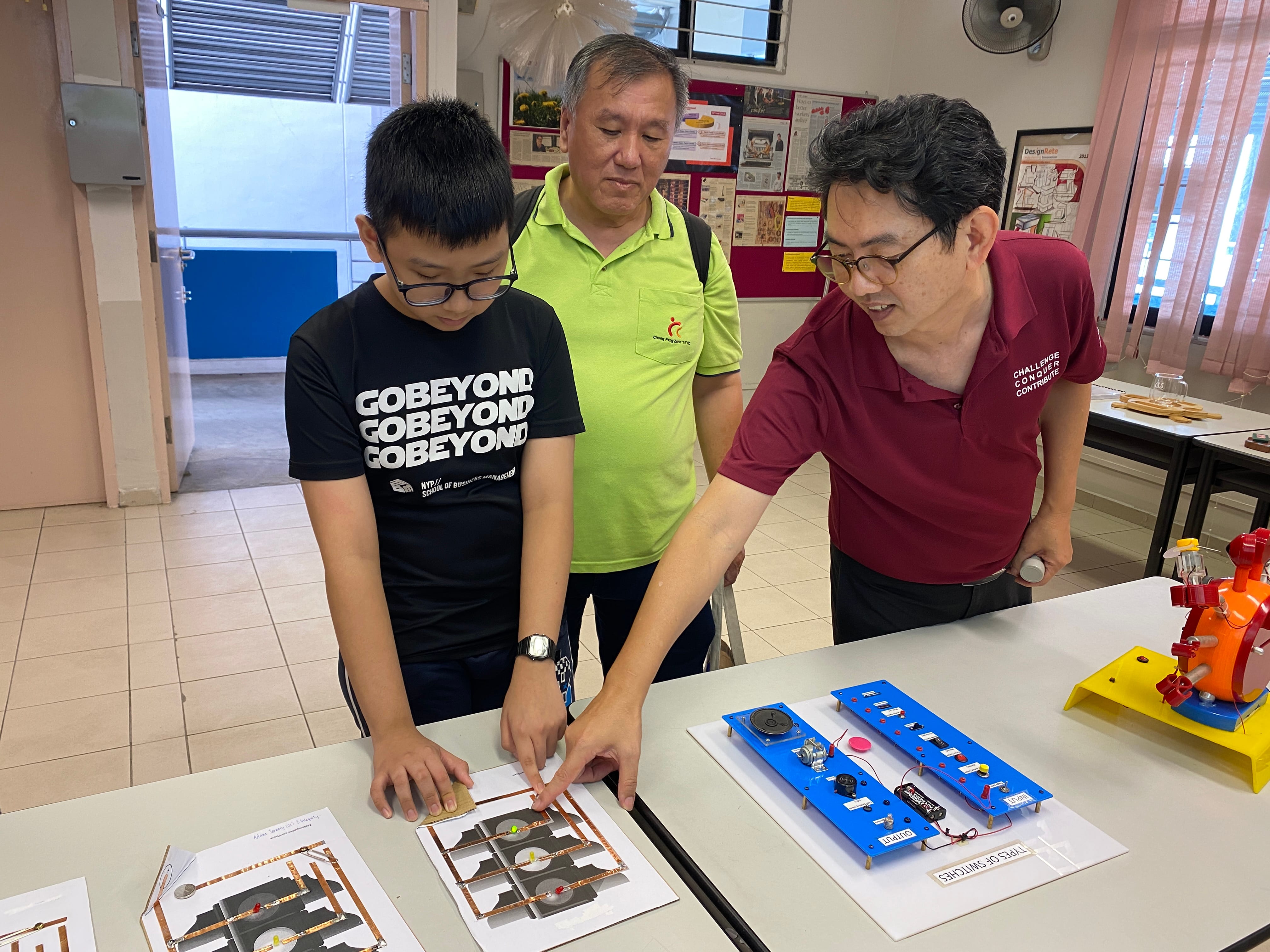 Two men and a boy examine circuit boards and electronics displays on a table in a classroom setting.