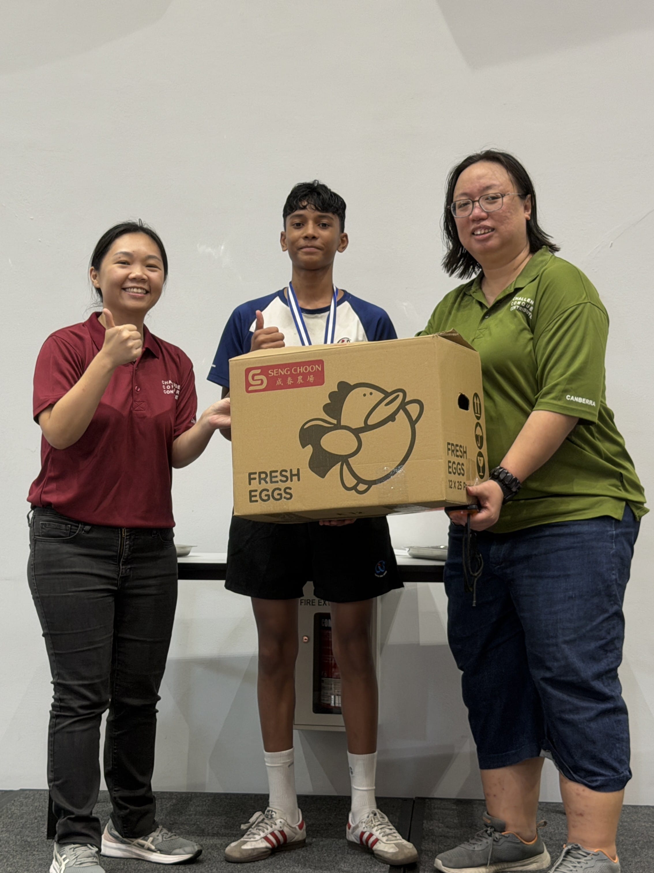 Two adults in maroon and green polo shirts flank a young male student wearing a medal, all three holding a Seng Choon Fresh Eggs cardboard box. Both adults give a thumbs up.