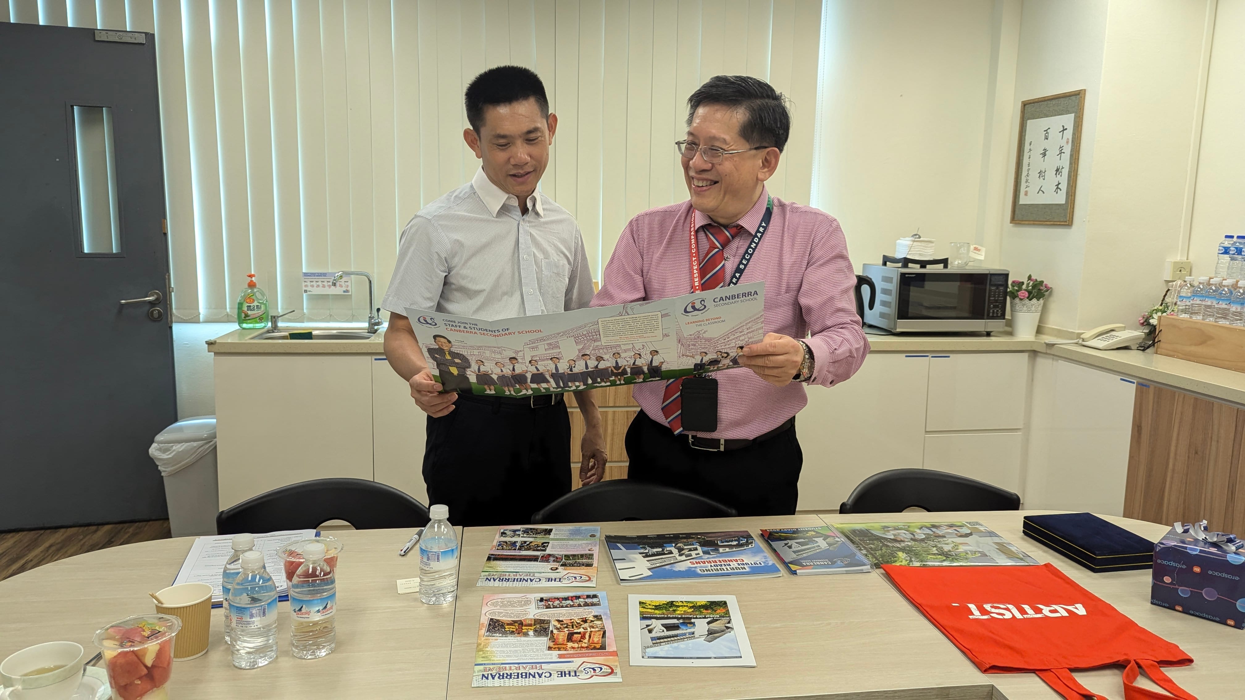 Two men stand together holding a Canberra Secondary School brochure, smiling, with pamphlets on a table before them.