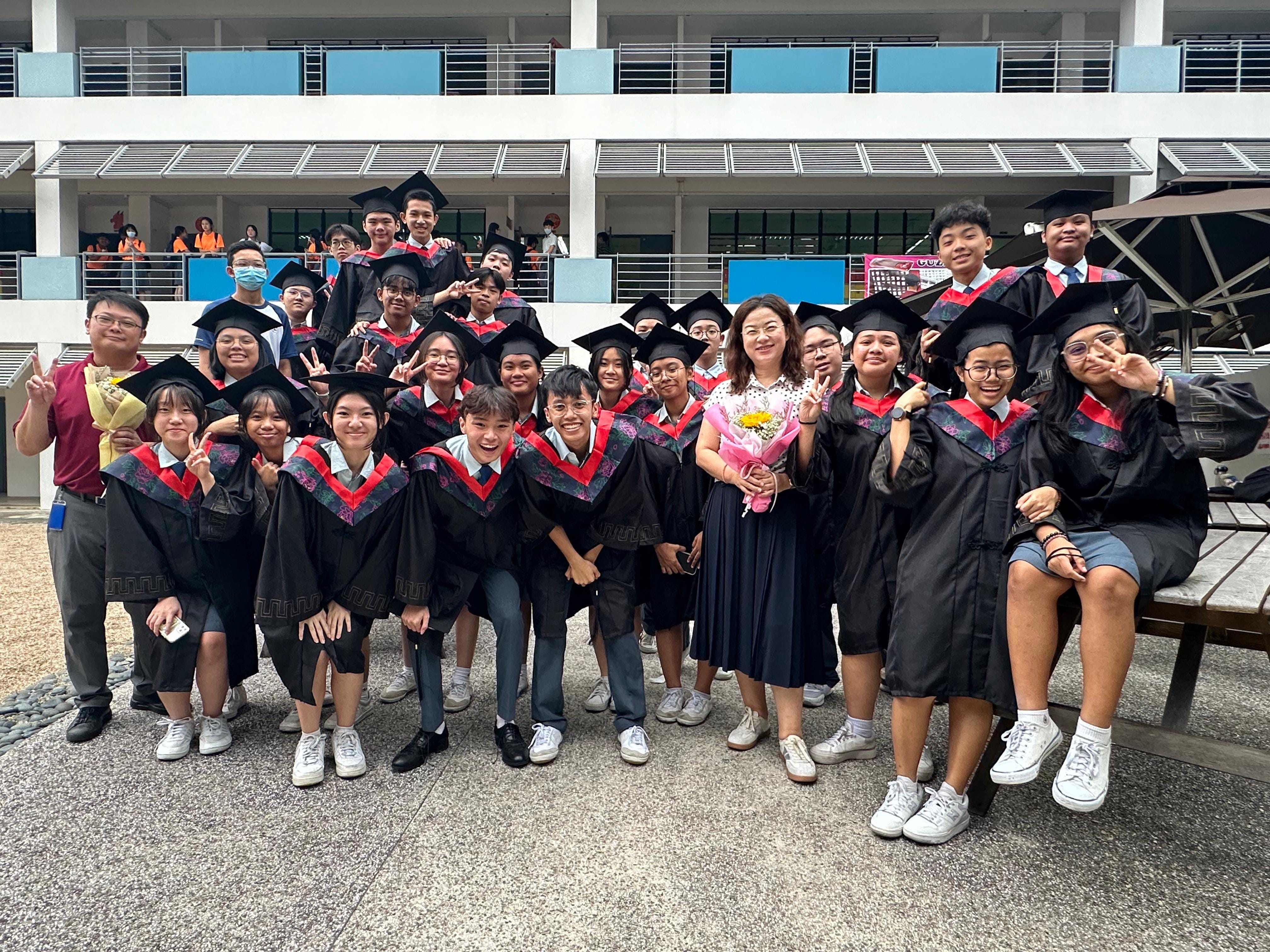 A large group of students in black graduation gowns and caps pose outdoors with a teacher holding flowers.